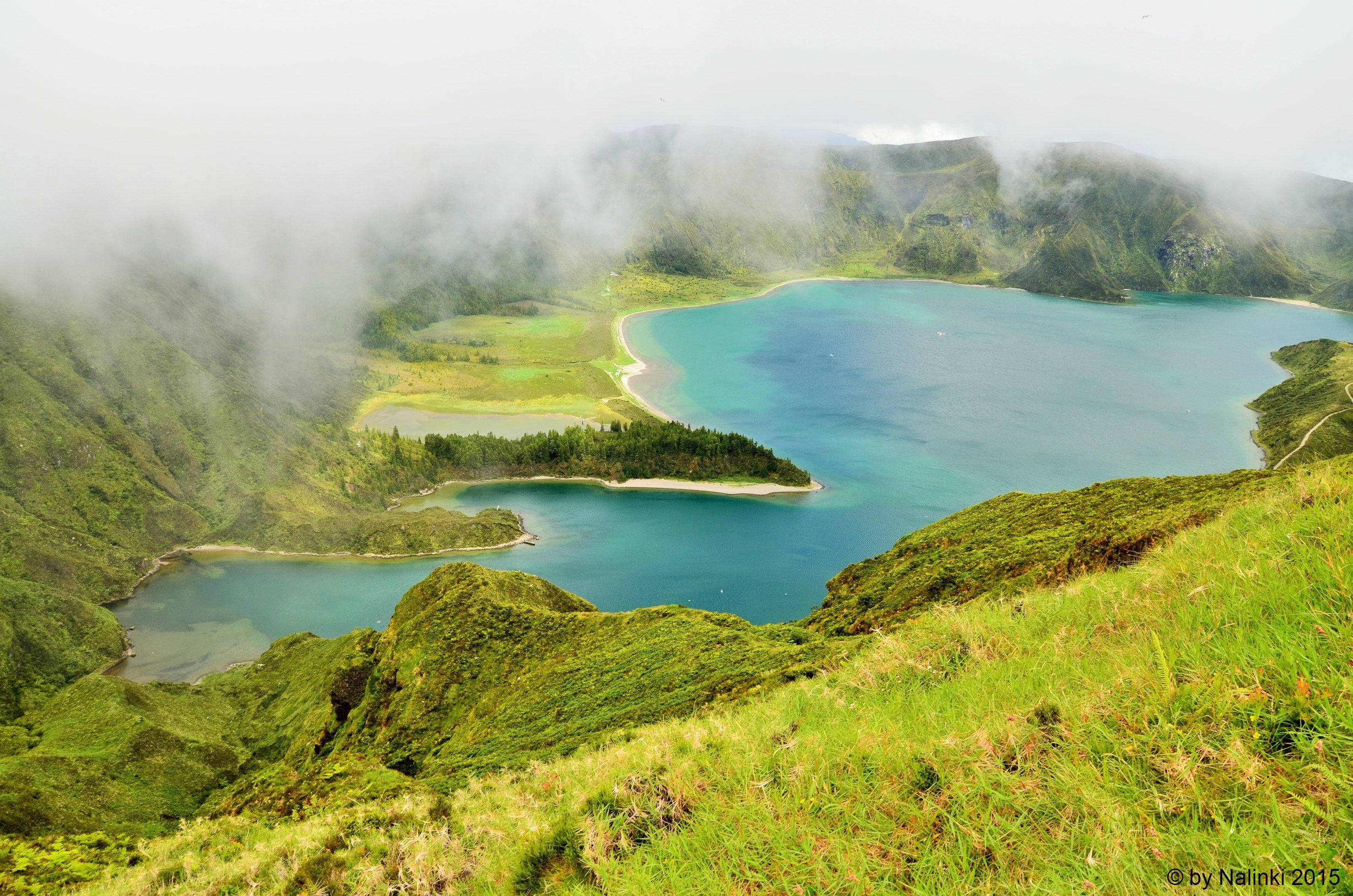 Portugal 桌布 壁紙 — 一個霧氣瀰漫的火山口湖，湖水呈綠松石色，坐落在鬱鬱蔥蔥的綠色山丘之間，天空多雲。