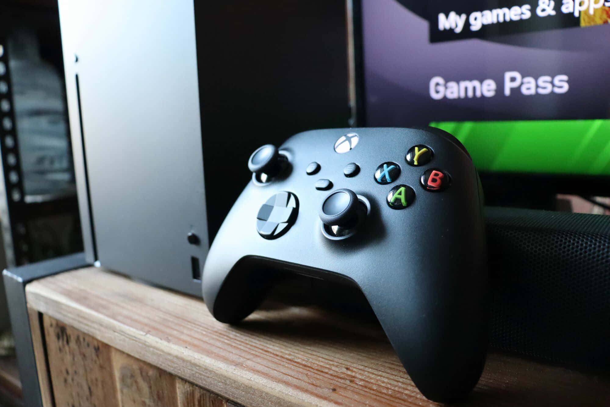 Xbox 桌布 壁紙 — 一個霧面黑色Xbox手把放在木質表面上，旁邊是遊戲主機和顯示Game Pass的電視。