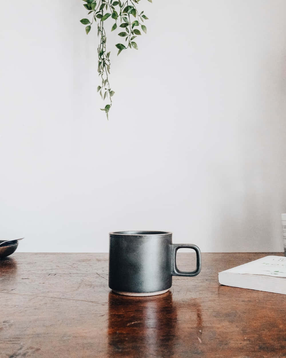 Minimalist Coffee 桌布 壁紙 — 一個霧面黑色馬克杯放在質樸木桌中央，旁邊有一本書和垂吊的綠色植物，背景是純白牆壁。