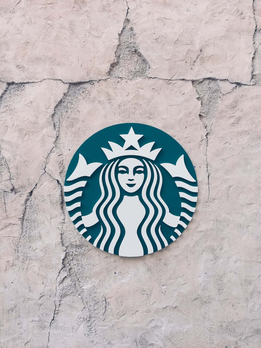 Aesthetic Starbucks 桌布 壁紙 — 一個青綠色和白色的星巴克海妖標誌安裝在裂縫的米色混凝土牆上。