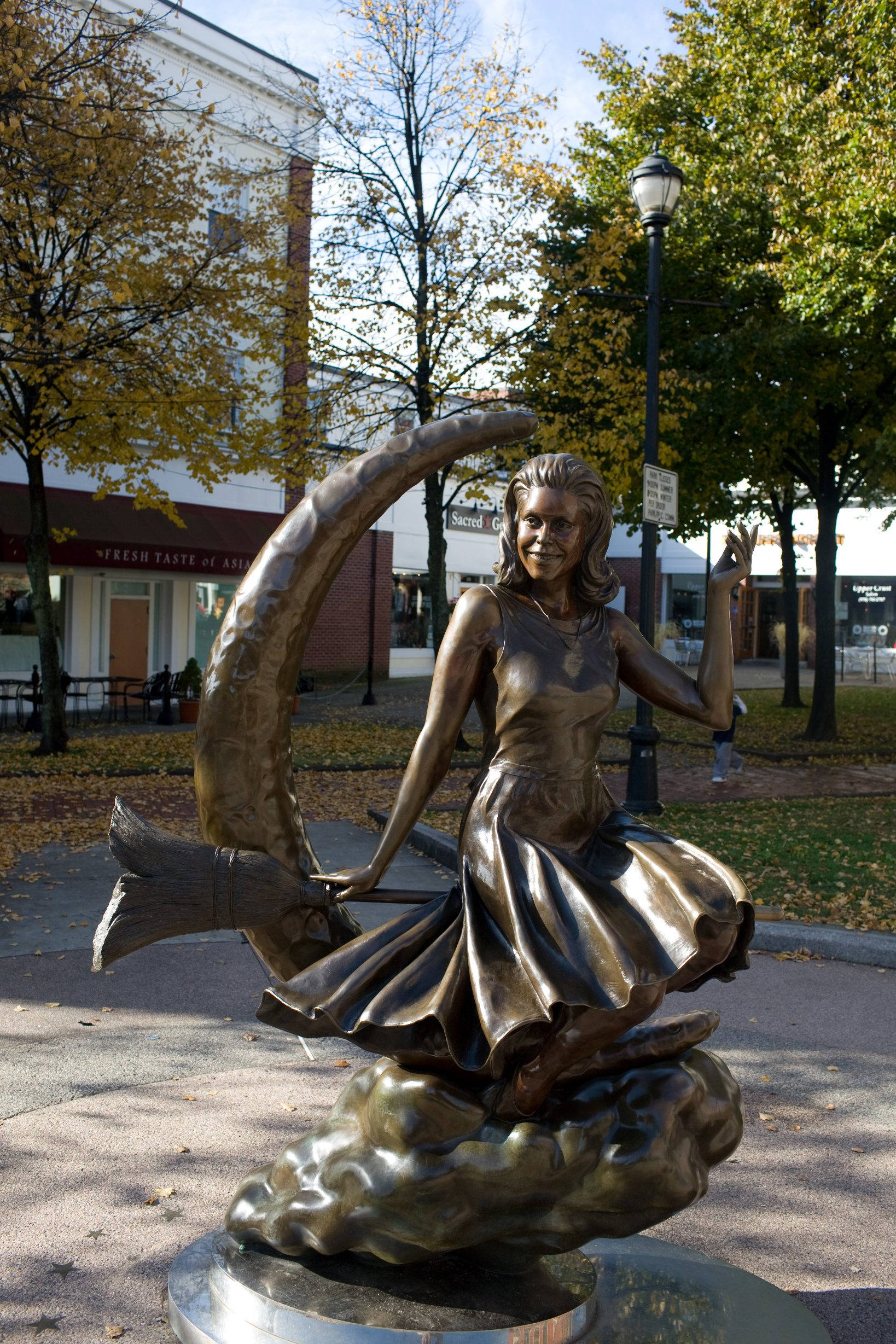 Statue Aesthetic 桌布 壁紙 — 一個青銅雕像，描繪一個女孩在城鎮廣場上跳舞，帶有新月和魚尾，周圍是秋天的樹木。