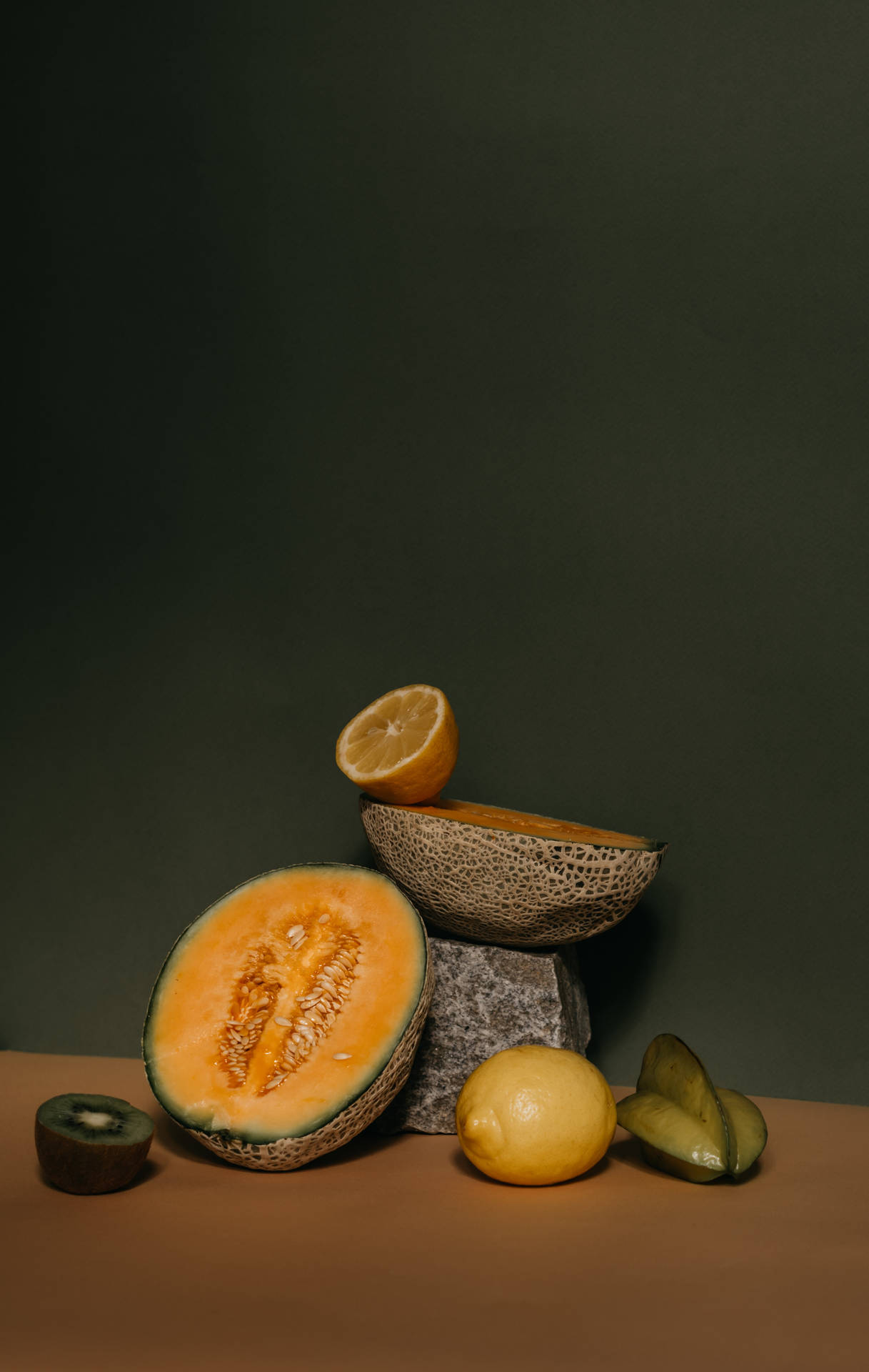 Fruits 桌布 壁紙 — 一個靜物，將切好的蜜瓜、奇異果、檸檬和楊桃放在石頭上，以深綠色牆壁為背景。