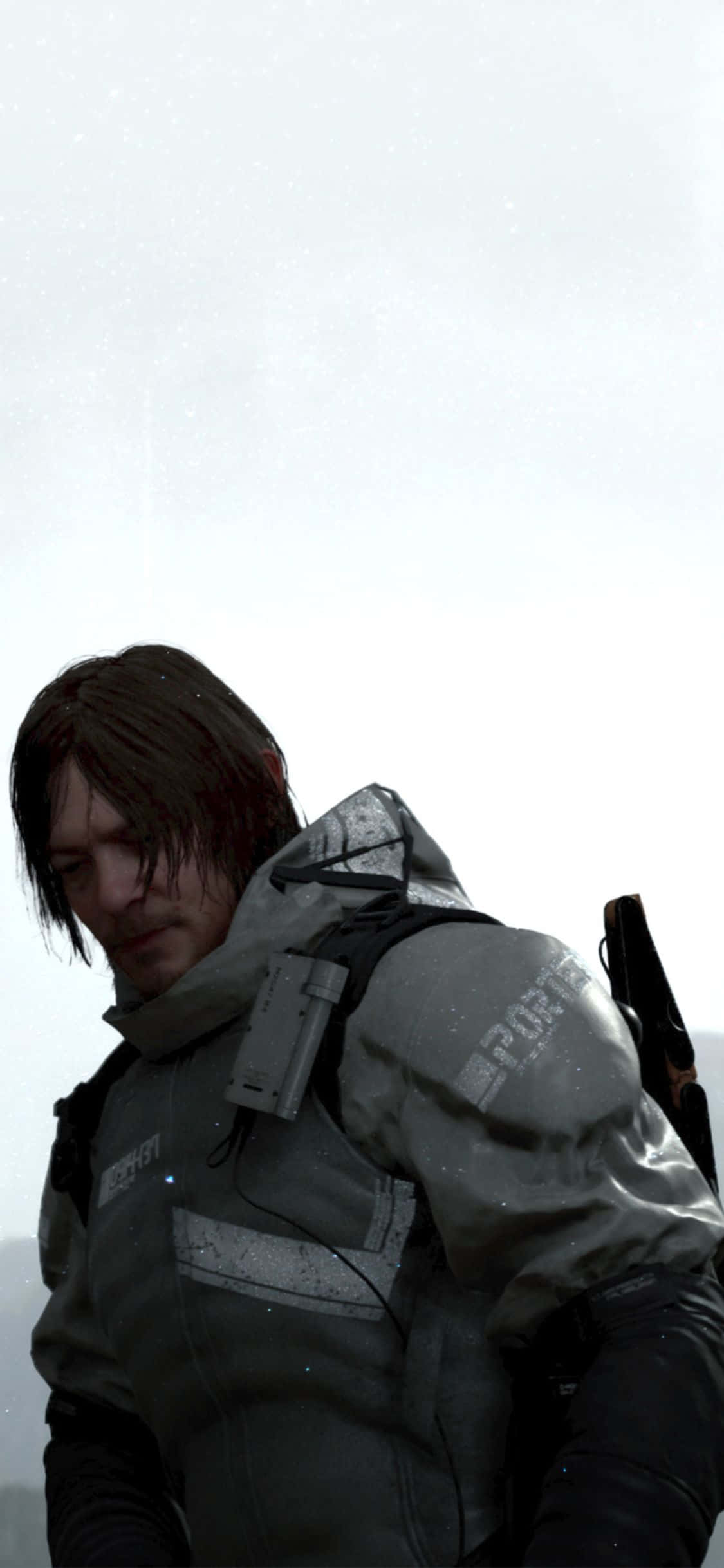 iPhone Xs Death Stranding 桌布 壁紙 — 一個面容嚴峻的男人穿著灰色戰術外套，站在雪地陰天的背景下，背上綁著武器。