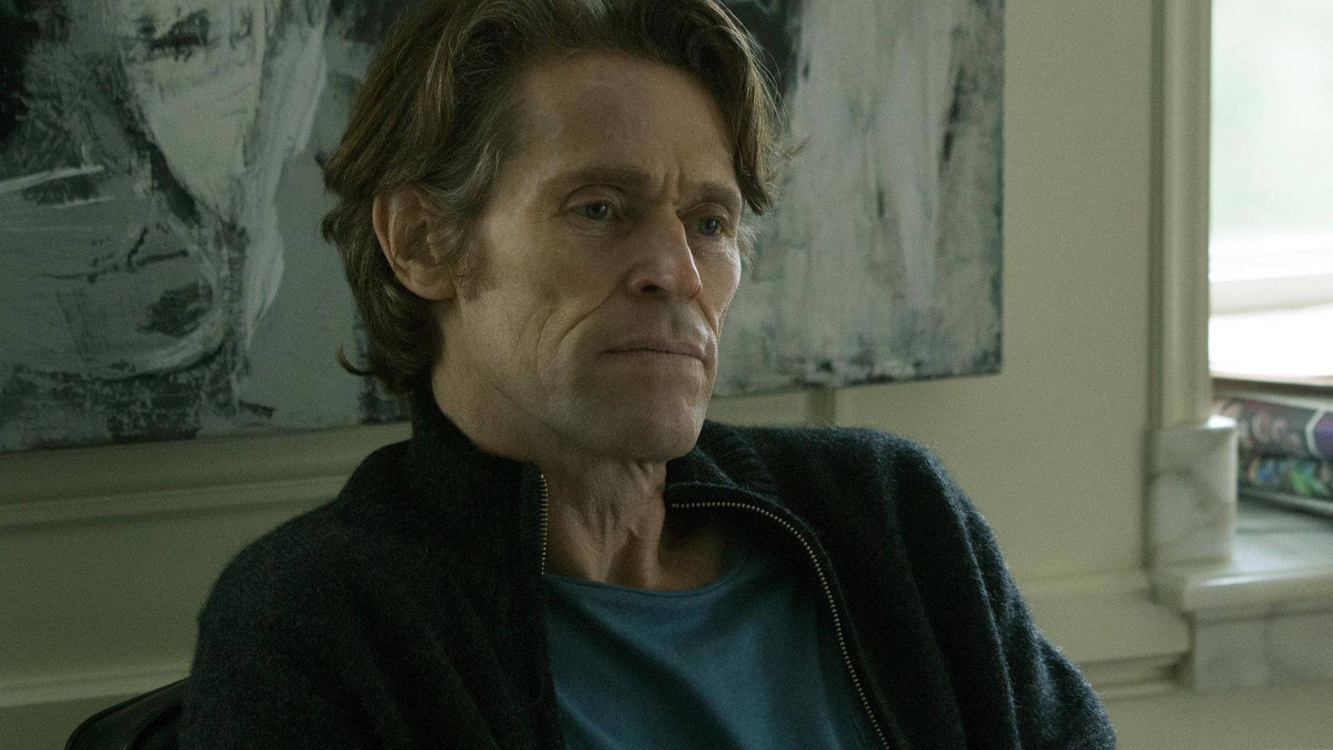 Willem Dafoe 桌布 壁紙 — 一個面容滄桑、眼神銳利的男人坐在光線柔和的房間裡，身後牆上掛著抽象藝術品。