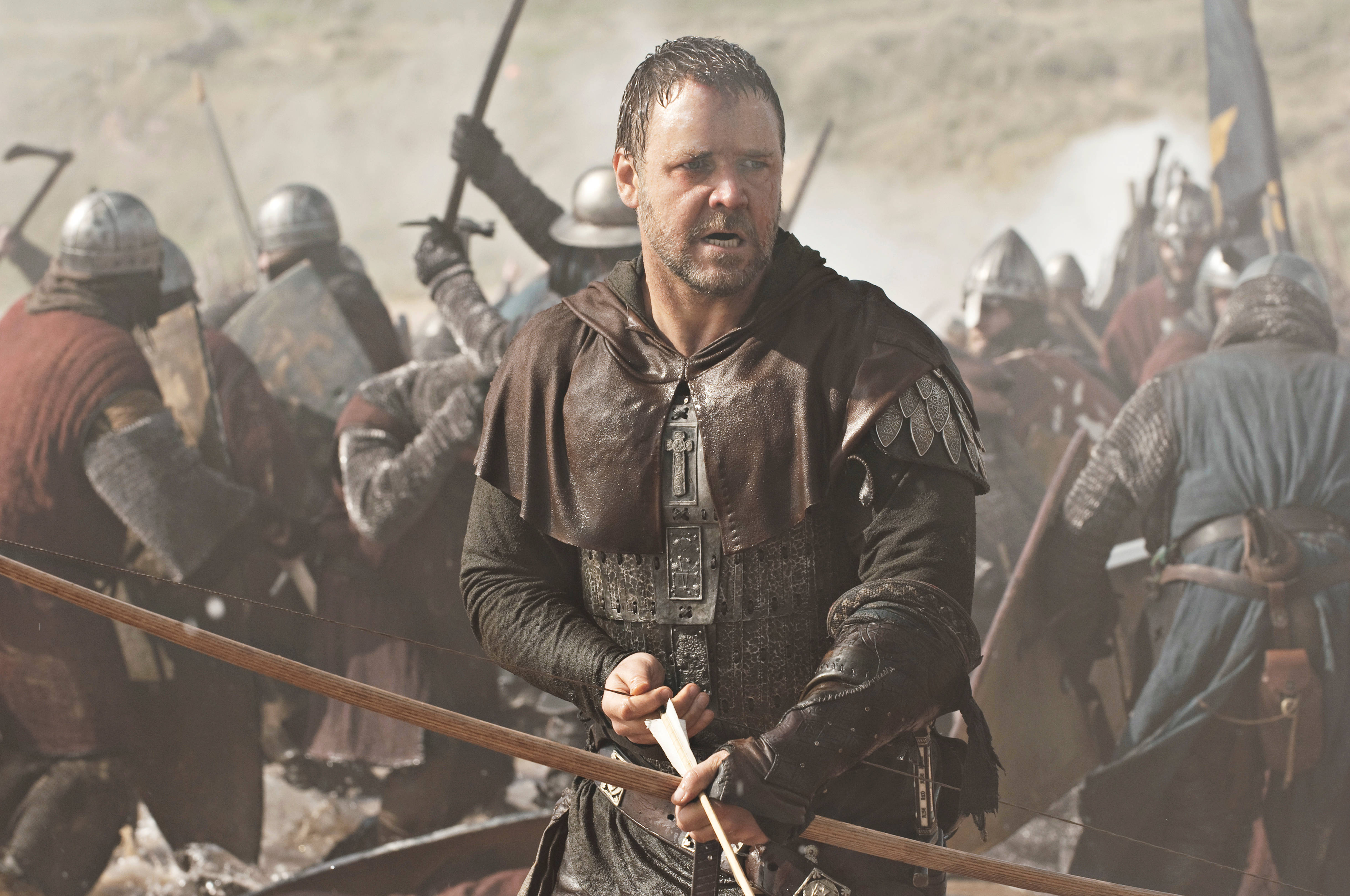 Russell Crowe 桌布 壁紙 — 一個面容陰沉的中世紀戰士身穿皮甲，在塵土飛揚的背景中，於混亂的戰場上握緊弓，裝甲士兵正在交戰。