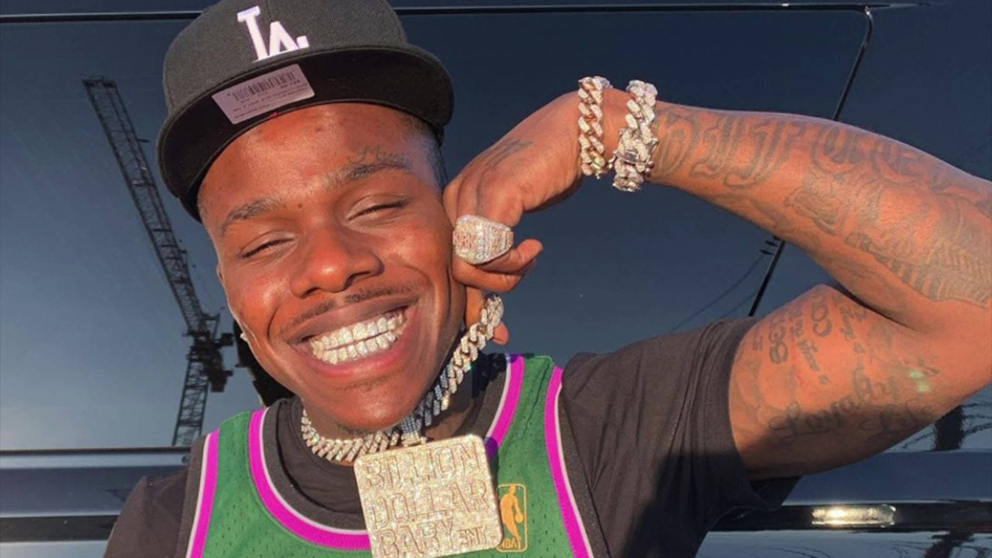 Dababy 桌布 壁紙 — 一個面帶微笑、有刺青和閃亮珠寶的男子在明亮的日光下，自豪地在一輛黑色汽車前擺姿勢。