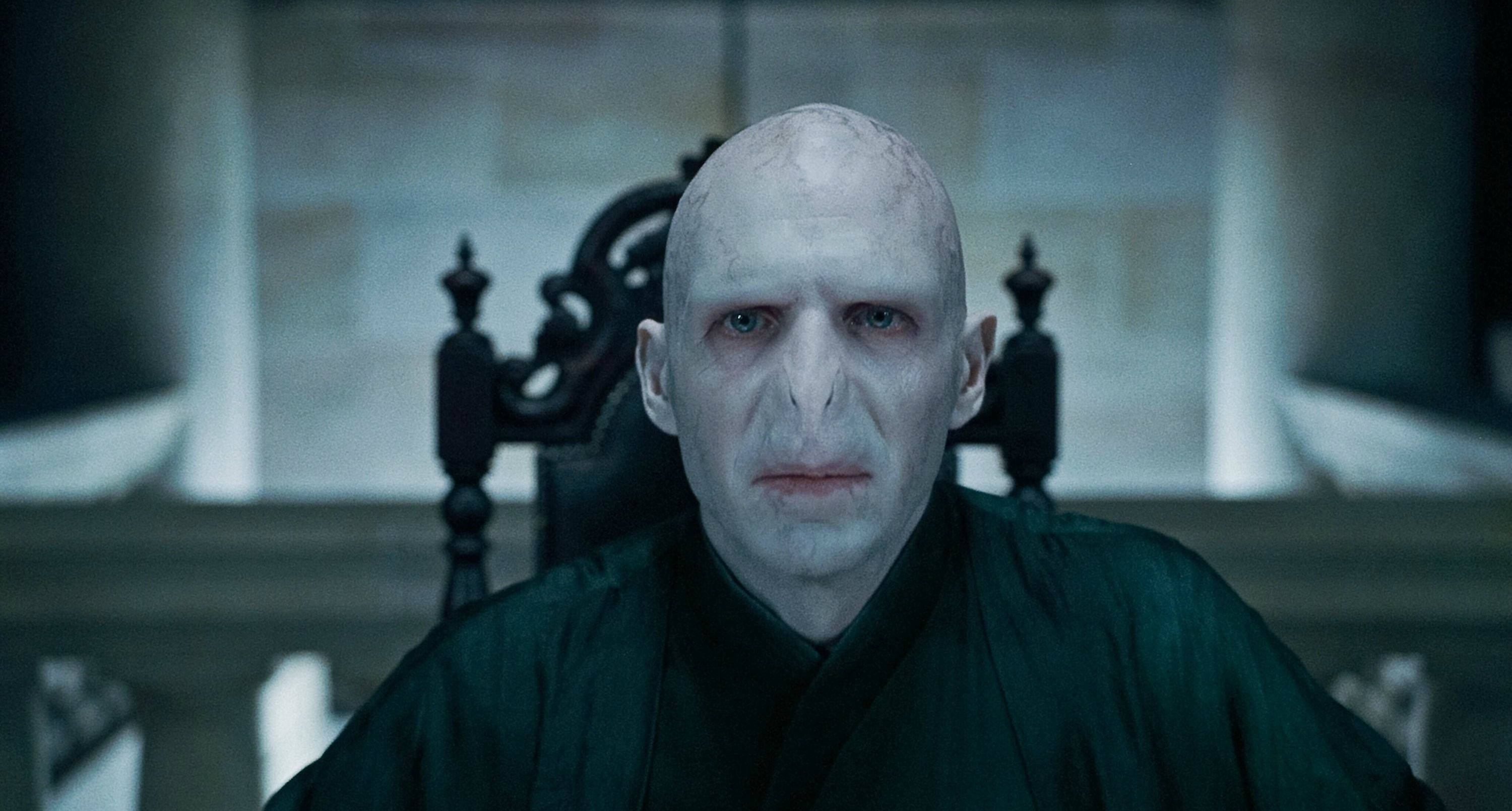 voldemort 桌布 壁紙 — 一個面色蒼白的光頭男人，紅眼睛，坐在昏暗石室裡一張深色雕花椅上。