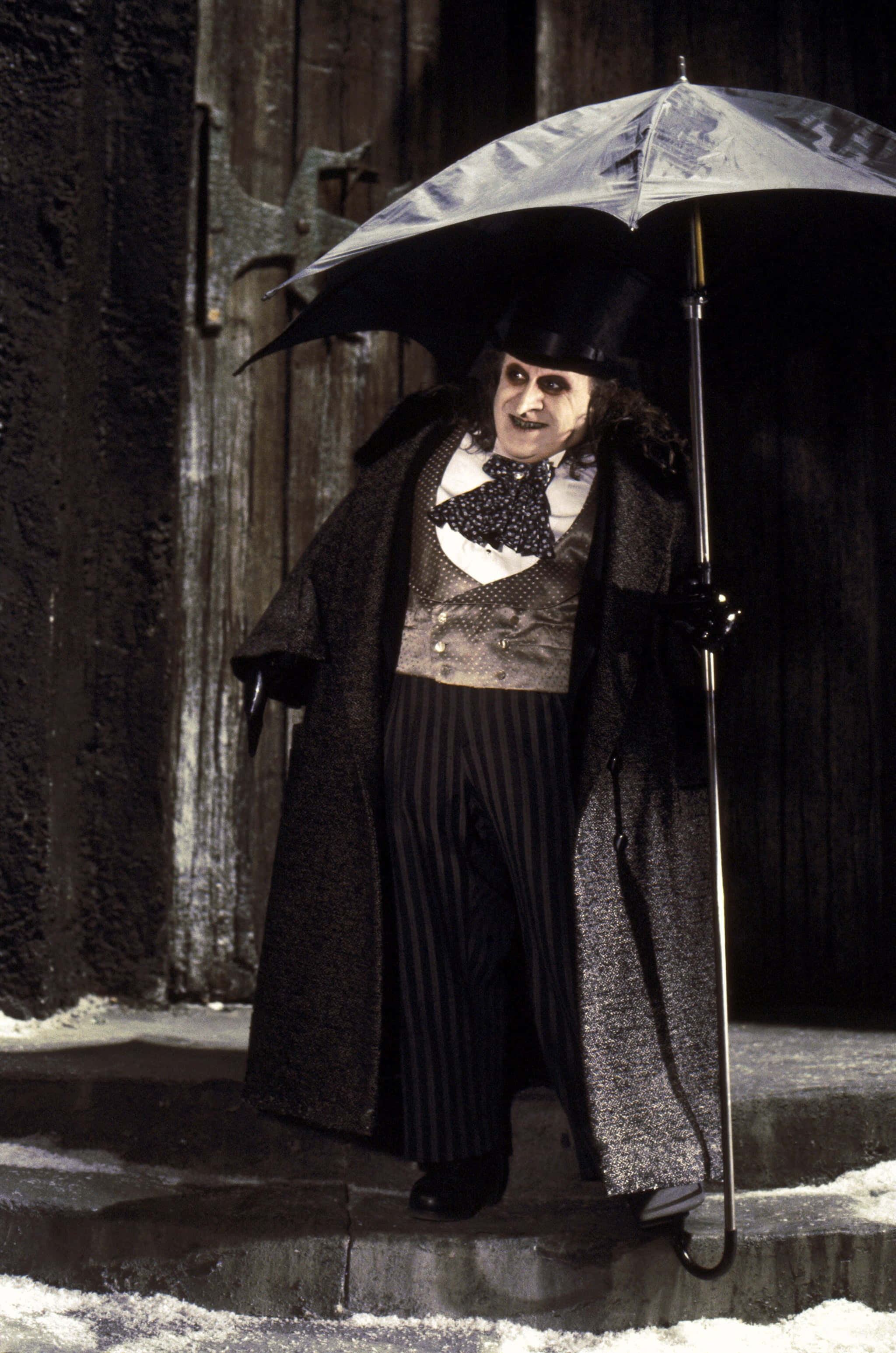 Batman Penguin 桌布 壁紙 — 一個面色蒼白的男人戴著高帽和長外套，站在雪白的階梯上，對著一扇深色木門，手持一把黑色雨傘。