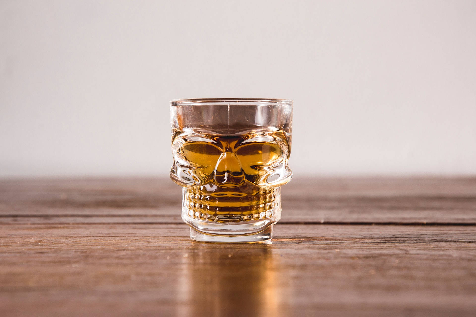 alcohol 桌布 壁紙 — 一個頭骨形狀的玻璃杯裝滿琥珀色液體，放在樸素背景前的鄉村風格木桌中央。