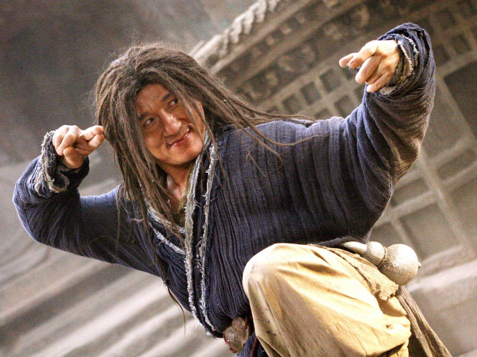 jackie chan 桌布 壁紙 — 一個頭髮凌亂、咧嘴而笑的男人，在模糊的石建築背景下擺出動態的武術姿勢。
