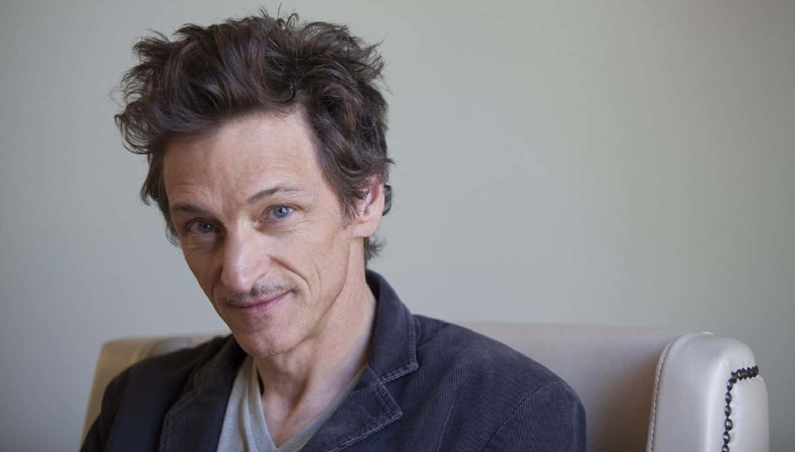 John Hawkes 桌布 壁紙 — 一個頭髮凌亂深色、帶著微妙笑容的男人坐在淺色椅子上，背景是素牆。
