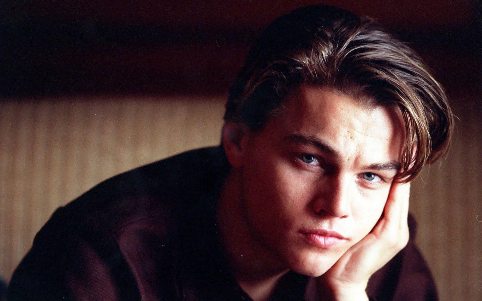 Dicaprio 桌布 壁紙 — 一個頭髮凌亂的年輕男子，手托下巴，在柔和光線的木質背景下凝視著鏡頭。