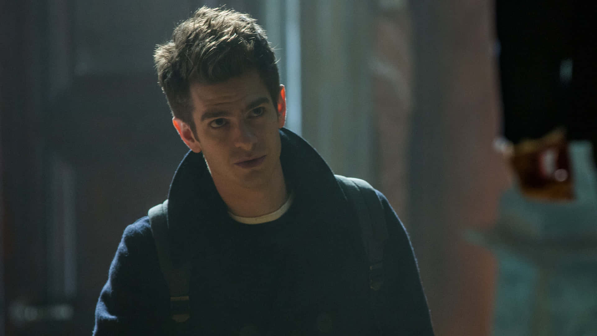 Andrew Garfield Spider Man 桌布 壁紙 — 一個頭髮凌亂的年輕男子在昏暗的室內燈光下穿著深色外套和背包。