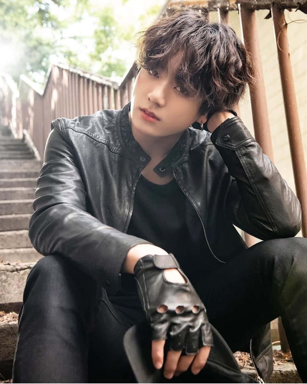 Jungkook 桌布 壁紙 — 一個頭髮凌亂的年輕男子穿著黑色皮夾克和露指手套坐在石階上，若有所思地向上凝視。