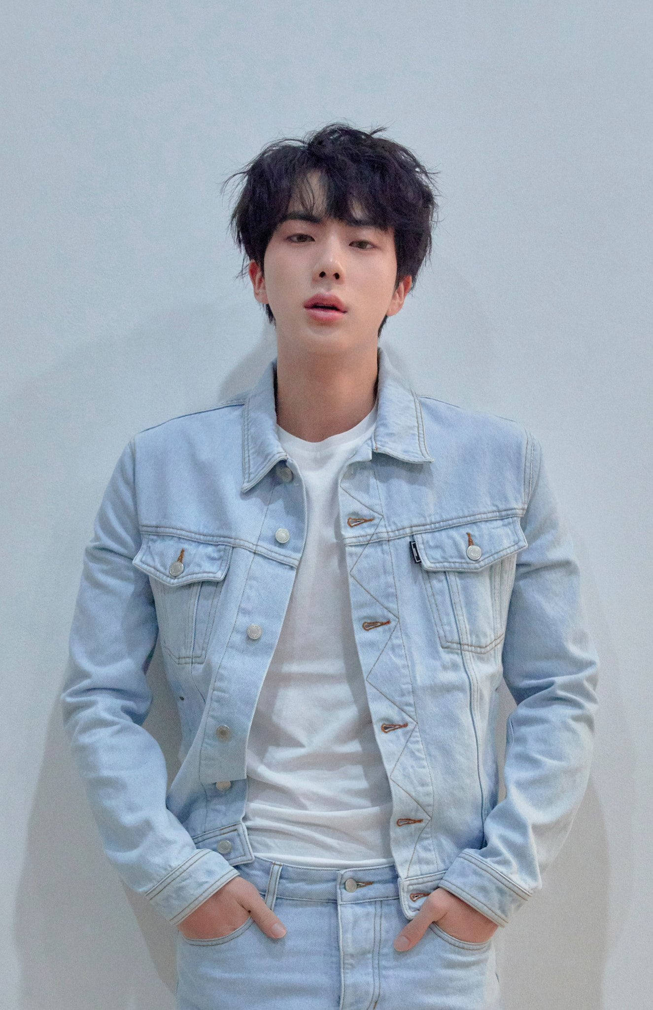 kim seok jin 桌布 壁紙 — 一個頭髮凌亂的深色頭髮年輕男子，穿著淺色牛仔外套和牛仔褲，站在純灰色牆壁前。