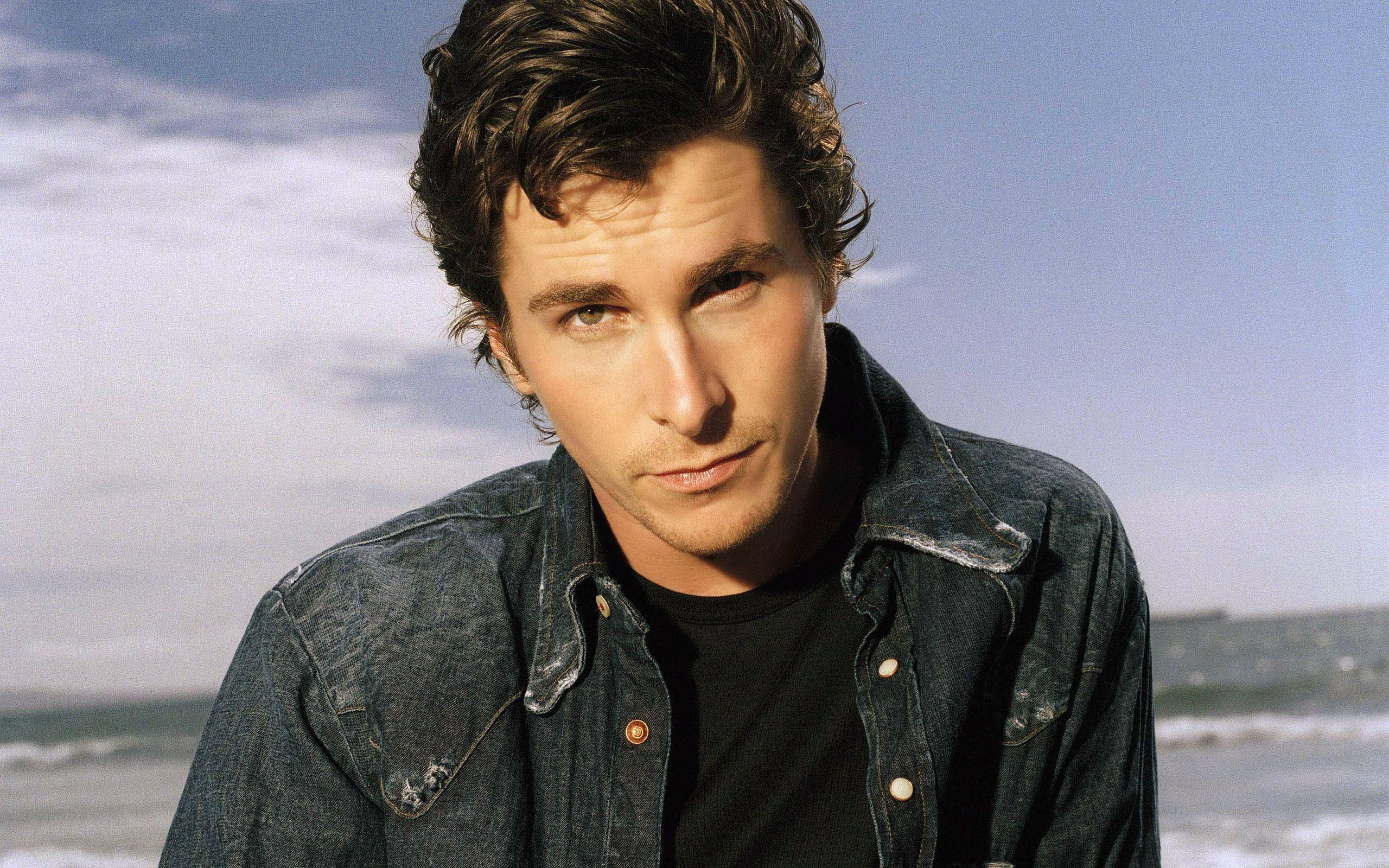 Christian Bale 桌布 壁紙 — 一個頭髮凌亂的深色頭髮男人穿著牛仔外套，在海洋波浪和陰天天空的背景下凝視著。
