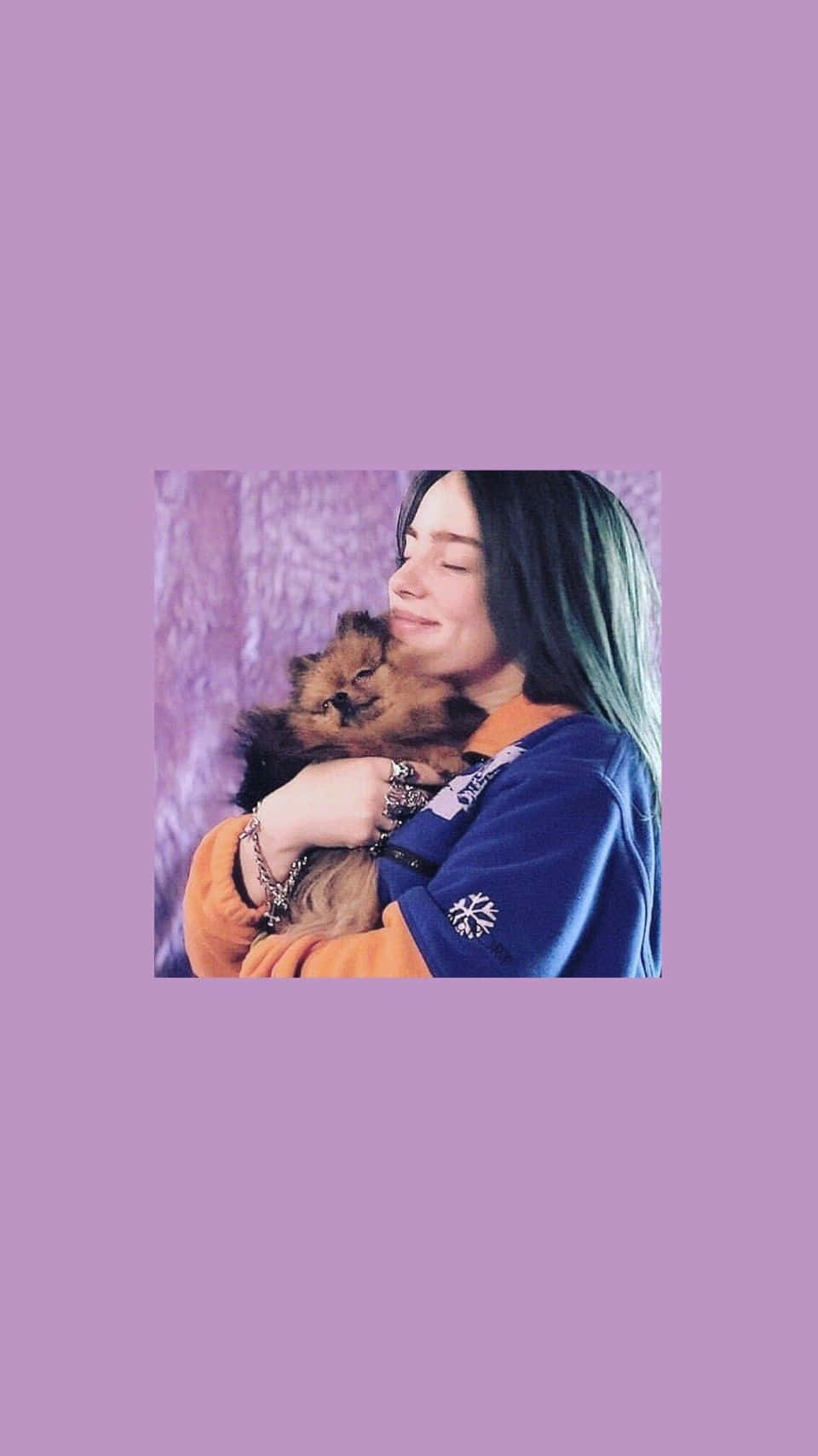 Billie Eilish Purple 桌布 壁紙 — 一個頭髮帶綠色尖端的人輕輕抱著一隻毛茸茸的棕色貓咪，背景是柔和的紫色。