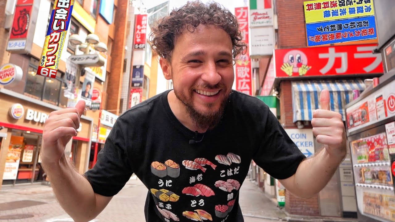 日本 桌布 壁紙 — 一個頭髮捲曲的開朗男子在繁華的日本街道上豎起兩個大拇指，街道兩旁是色彩繽紛的招牌和商店。