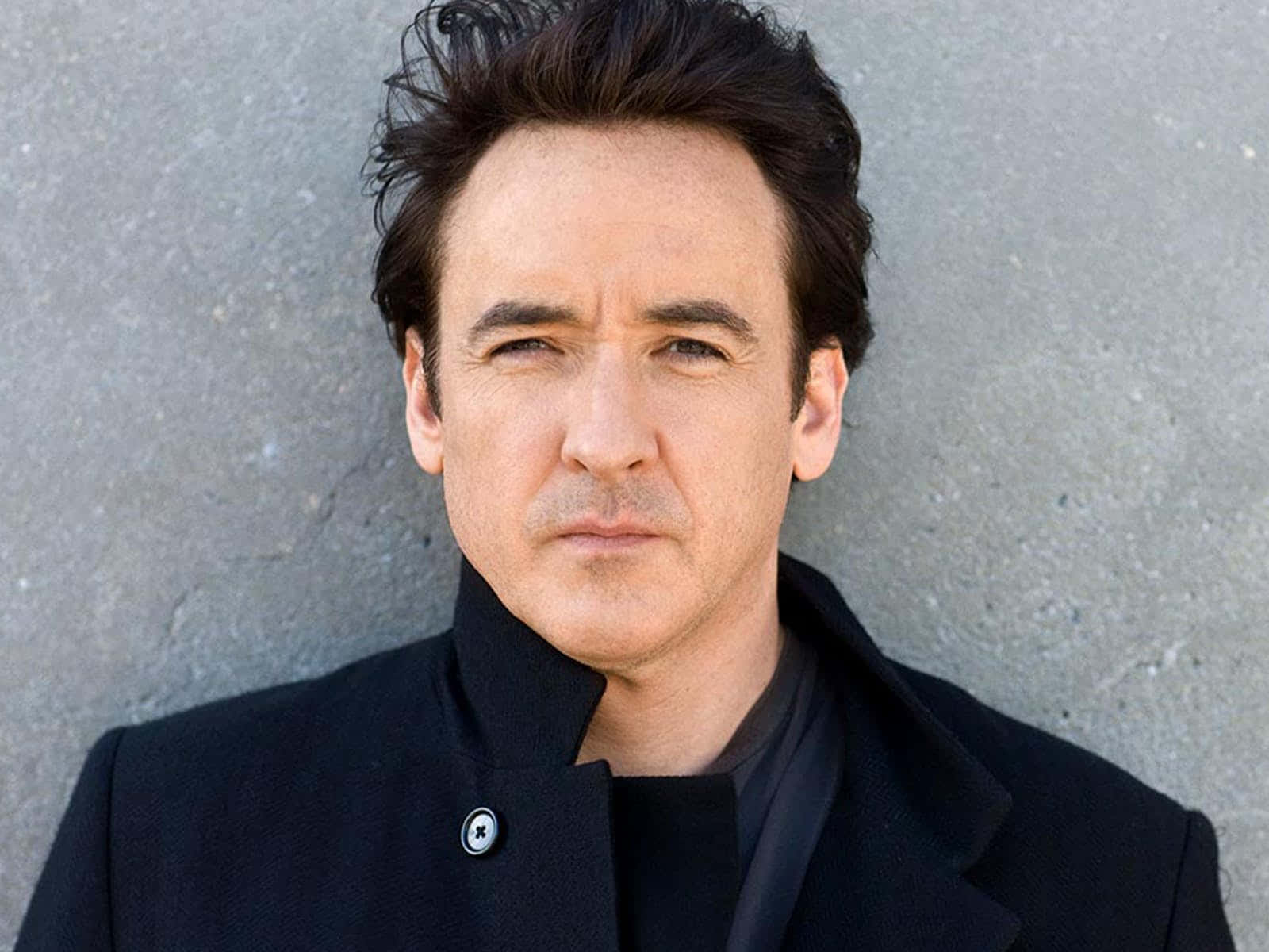 John Cusack 桌布 壁紙 — 一個頭髮深色凌亂、表情嚴肅的男人，身穿黑色外套，站在灰色混凝土牆前。