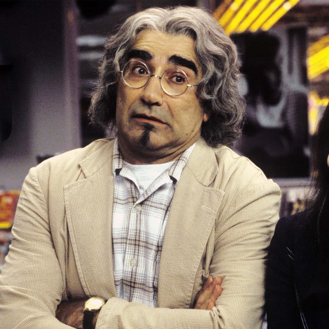 Eugene Levy 桌布 壁紙 — 一個頭髮灰白凌亂、戴著圓框眼鏡的男人，在溫暖的室內環境中睜大眼睛、交叉雙臂凝視著。