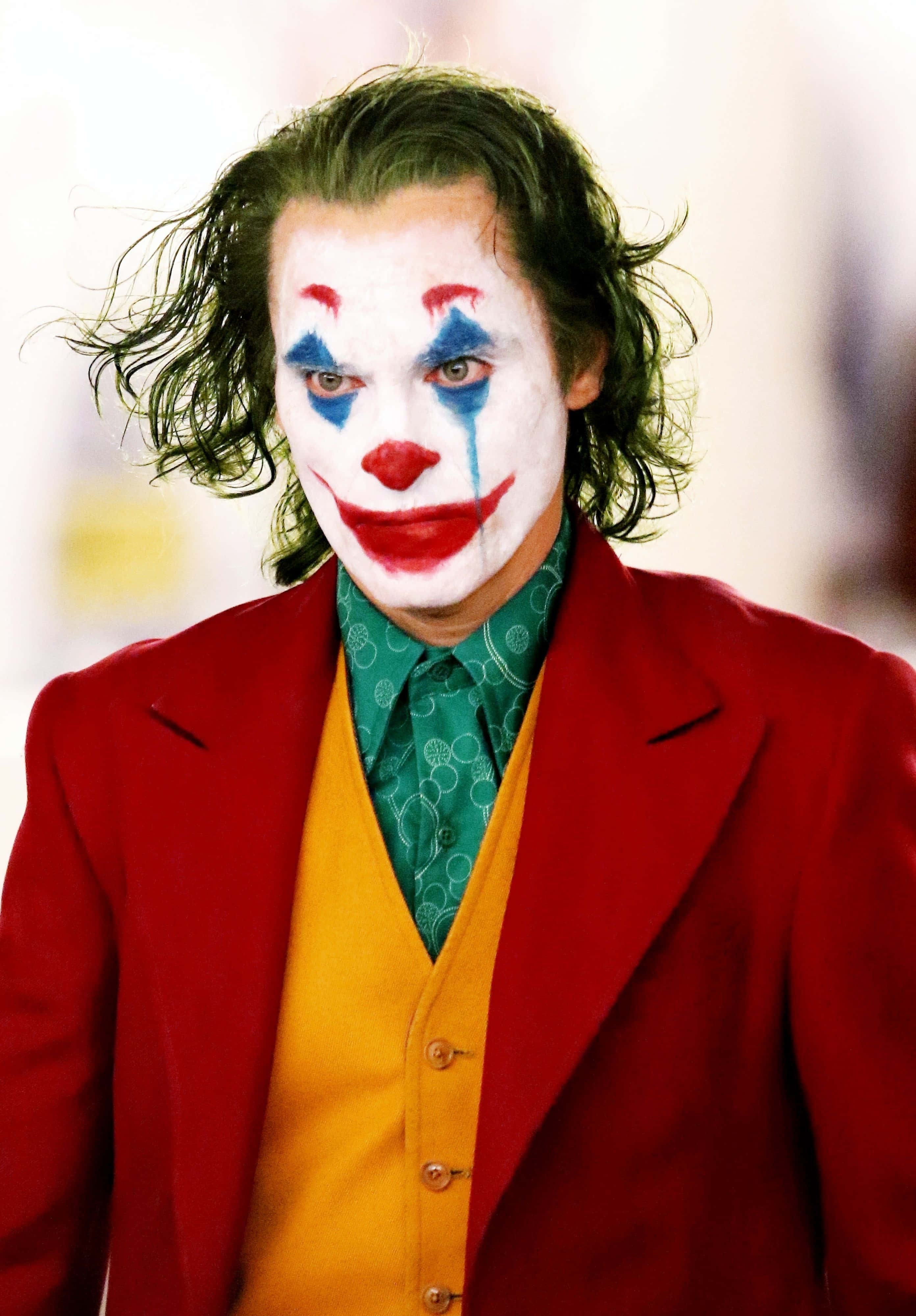 clown makeup 桌布 壁紙 — 一個頭髮狂野、化著小丑妝的男人，身穿紅色西裝和黃色背心，專注地凝視著鏡頭。