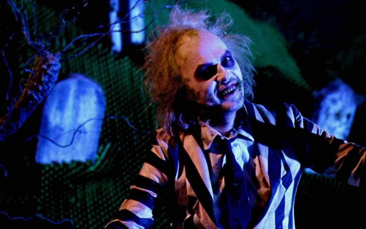Beetlejuice 桌布 壁紙 — 一個頭髮狂野、身穿條紋西裝的男人在陰森藍光下的黑暗墓園中狂笑。