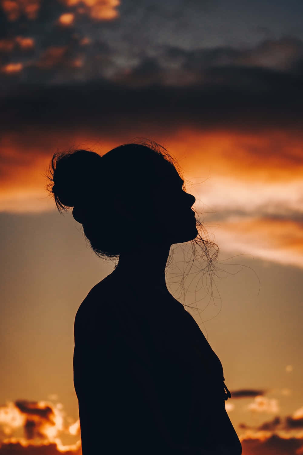Silhouette 桌布 壁紙 — 一個頭髮盤成髮髻的女人的剪影，在熾熱的橙色和紫色日落天空下向上凝望。