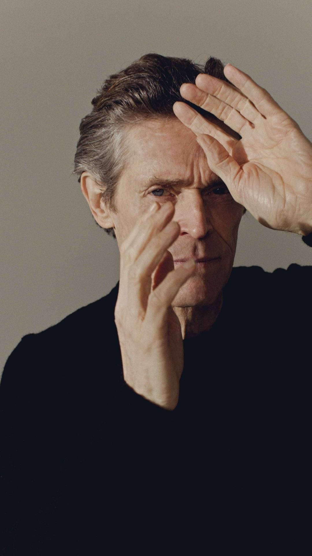 Willem Dafoe 桌布 壁紙 — 一個頭髮花白、眼神銳利的男人，雙手放在臉上，背景是純灰色。