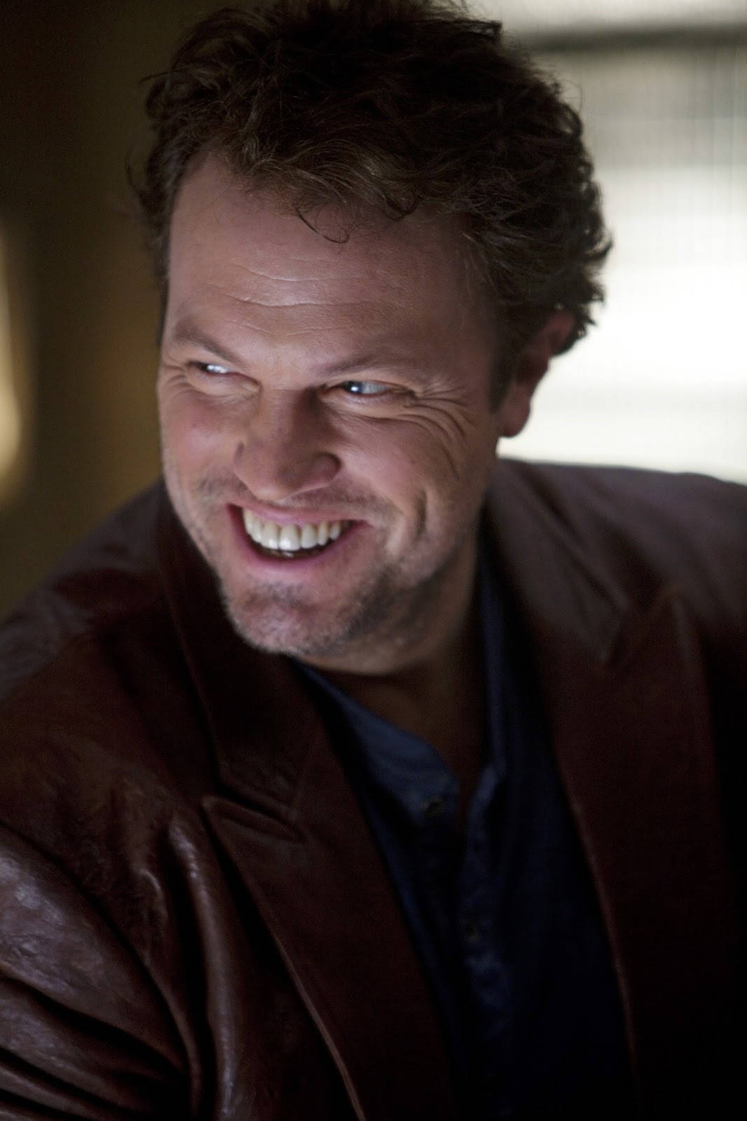 adam baldwin 桌布 壁紙 — 一個頭髮蓬鬆、咧嘴大笑的男人，在溫暖燈光的室內環境中穿著棕色皮夾克。