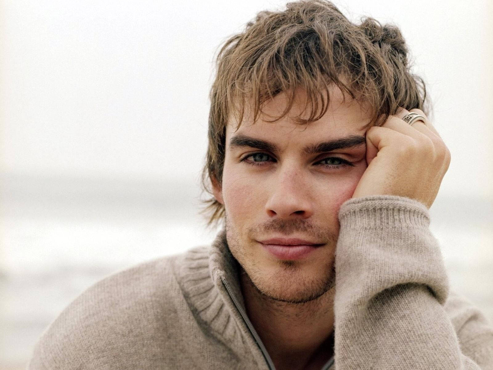 Ian Somerhalder 桌布 壁紙 — 一個頭髮蓬鬆、帶著溫柔微笑的男人，在柔和模糊的海灘背景下，將頭靠在手上。