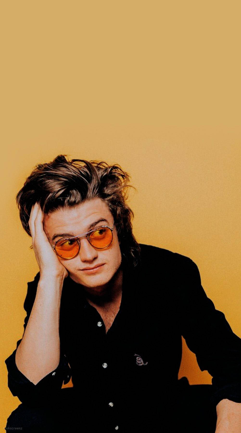 Joe Keery 桌布 壁紙 — 一個頭髮蓬鬆、戴著橙色眼鏡的年輕男子，在純芥末黃色背景上用手托著頭。