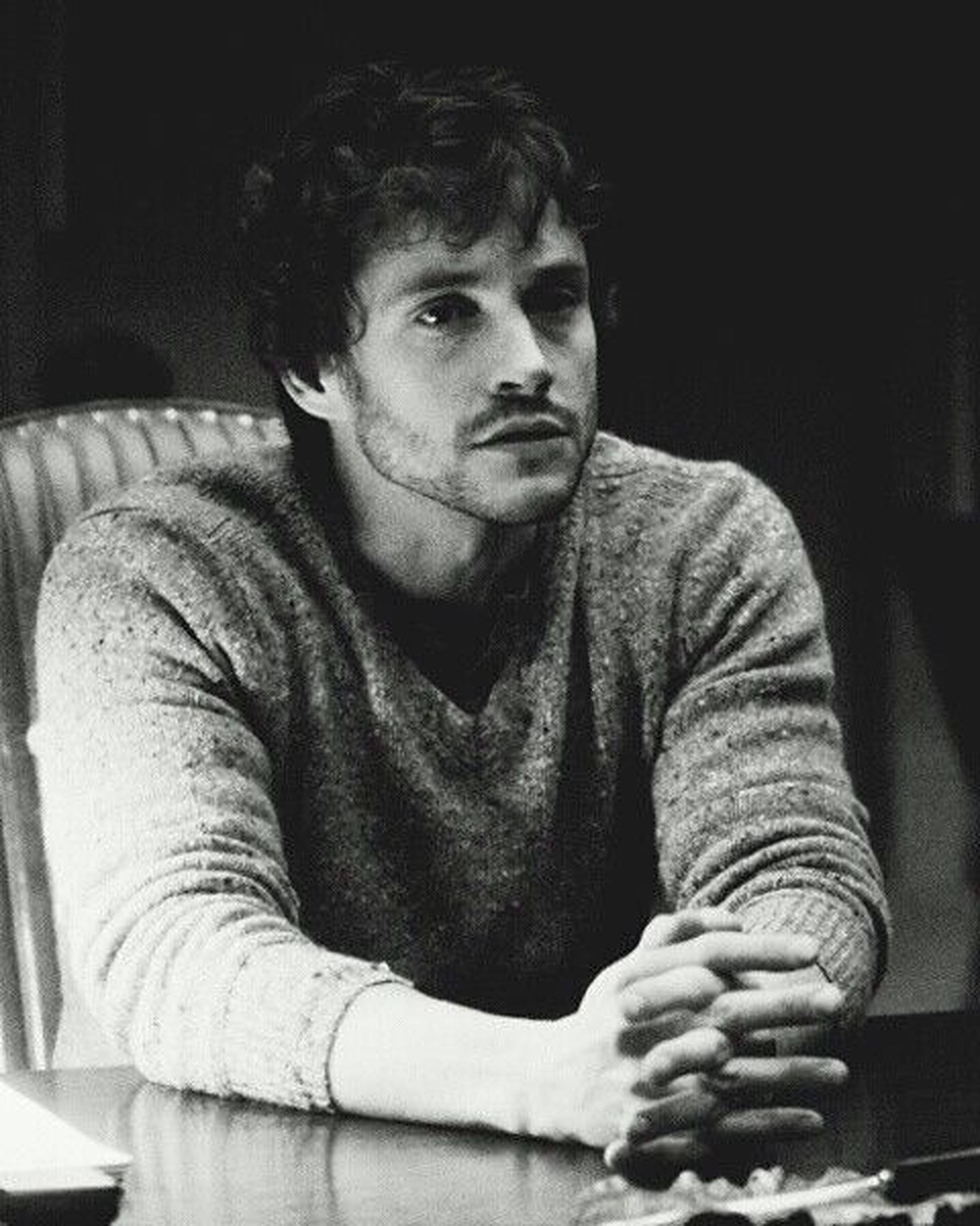 Hugh Dancy 桌布 壁紙 — 一個頭髮蓬鬆、留鬍子的男人坐在桌子旁，雙手緊握，一幅沉思的黑白肖像。