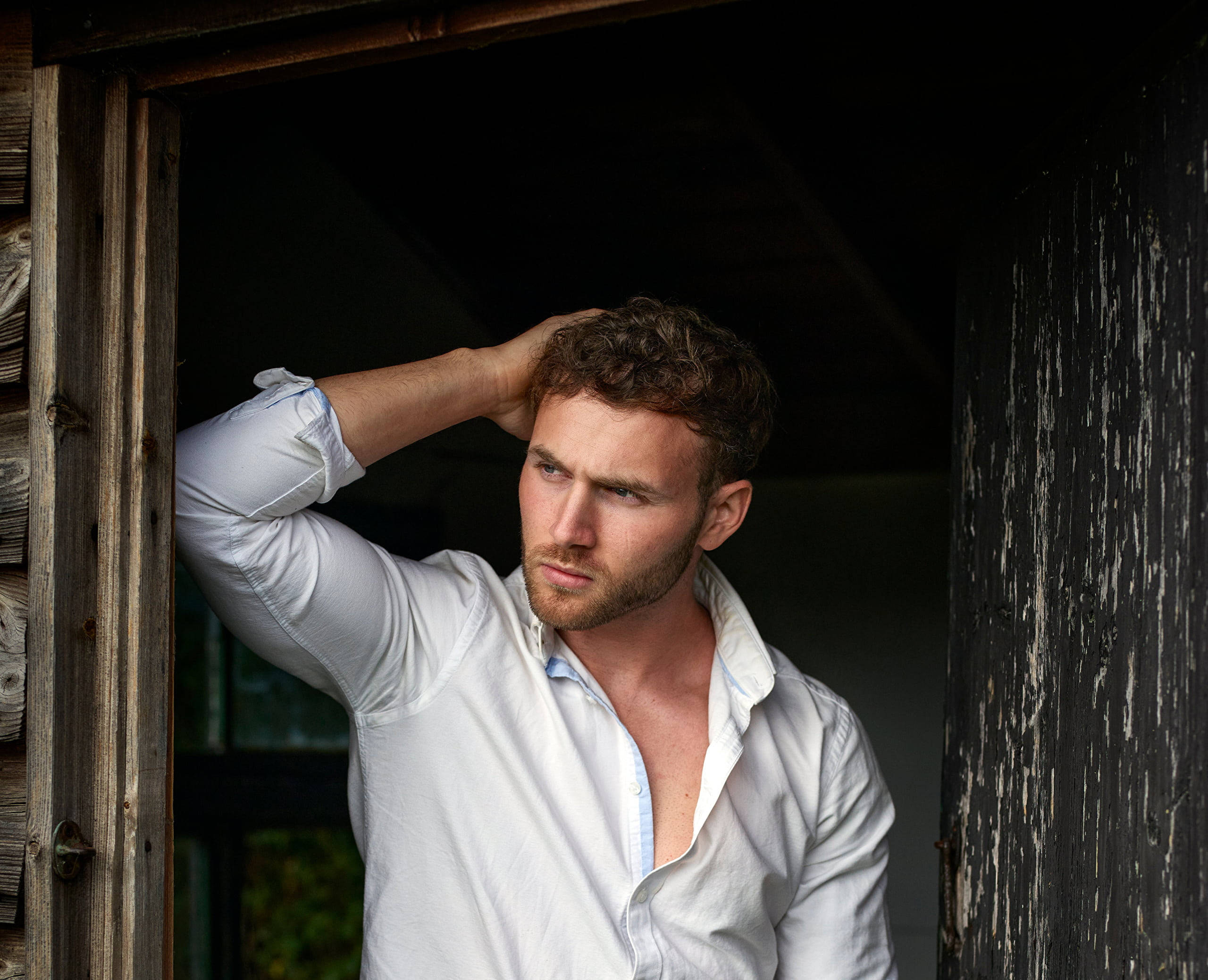 sexy man 桌布 壁紙 — 一個頭髮蓬鬆、留鬍鬚的男人站在質樸的木門廊中，手放在頭上，穿著一件敞開的白襯衫。
