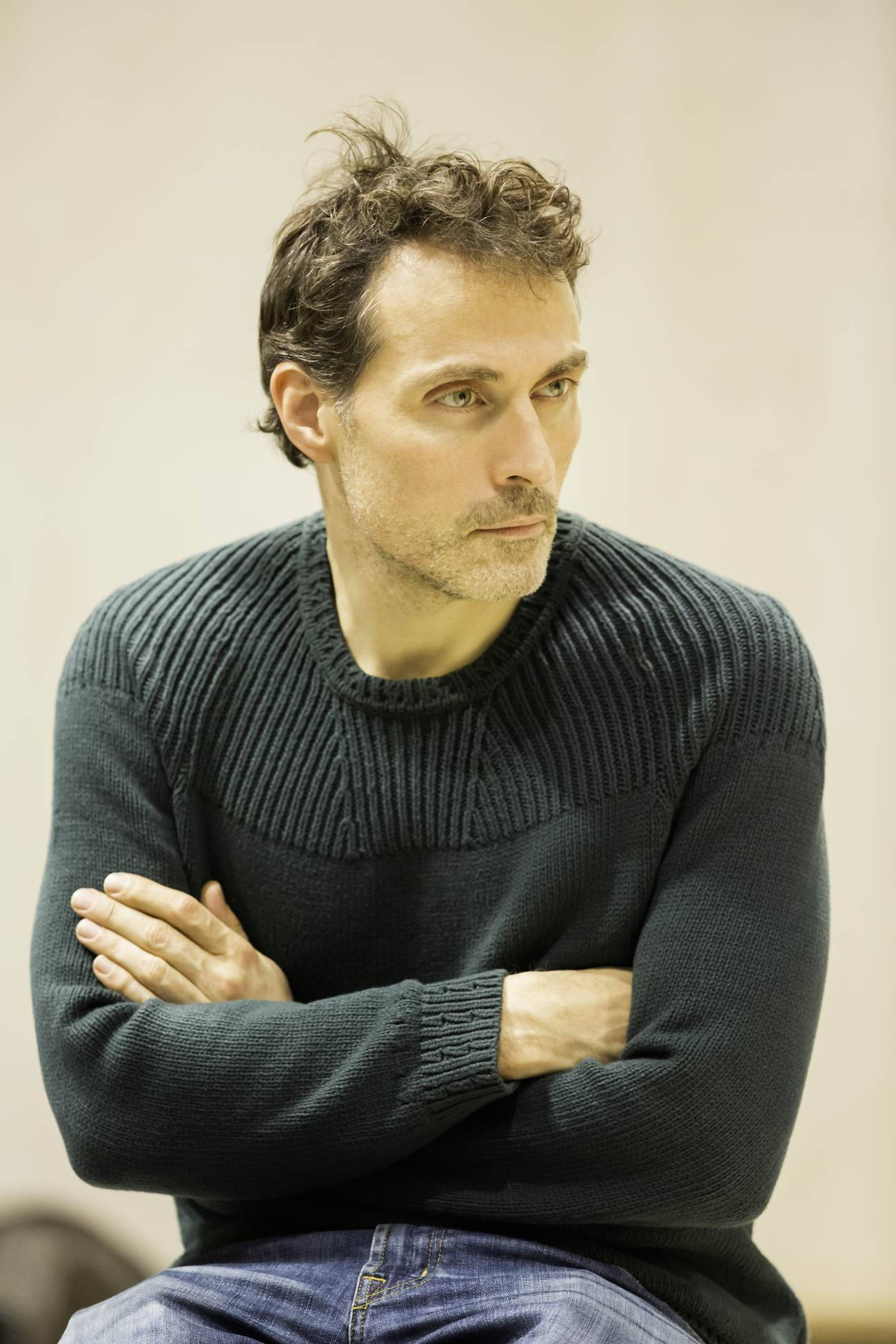 actor 桌布 壁紙 — 一個頭髮蓬鬆、身穿深色羅紋毛衣的男人，雙臂交叉，坐在純米色背景前。