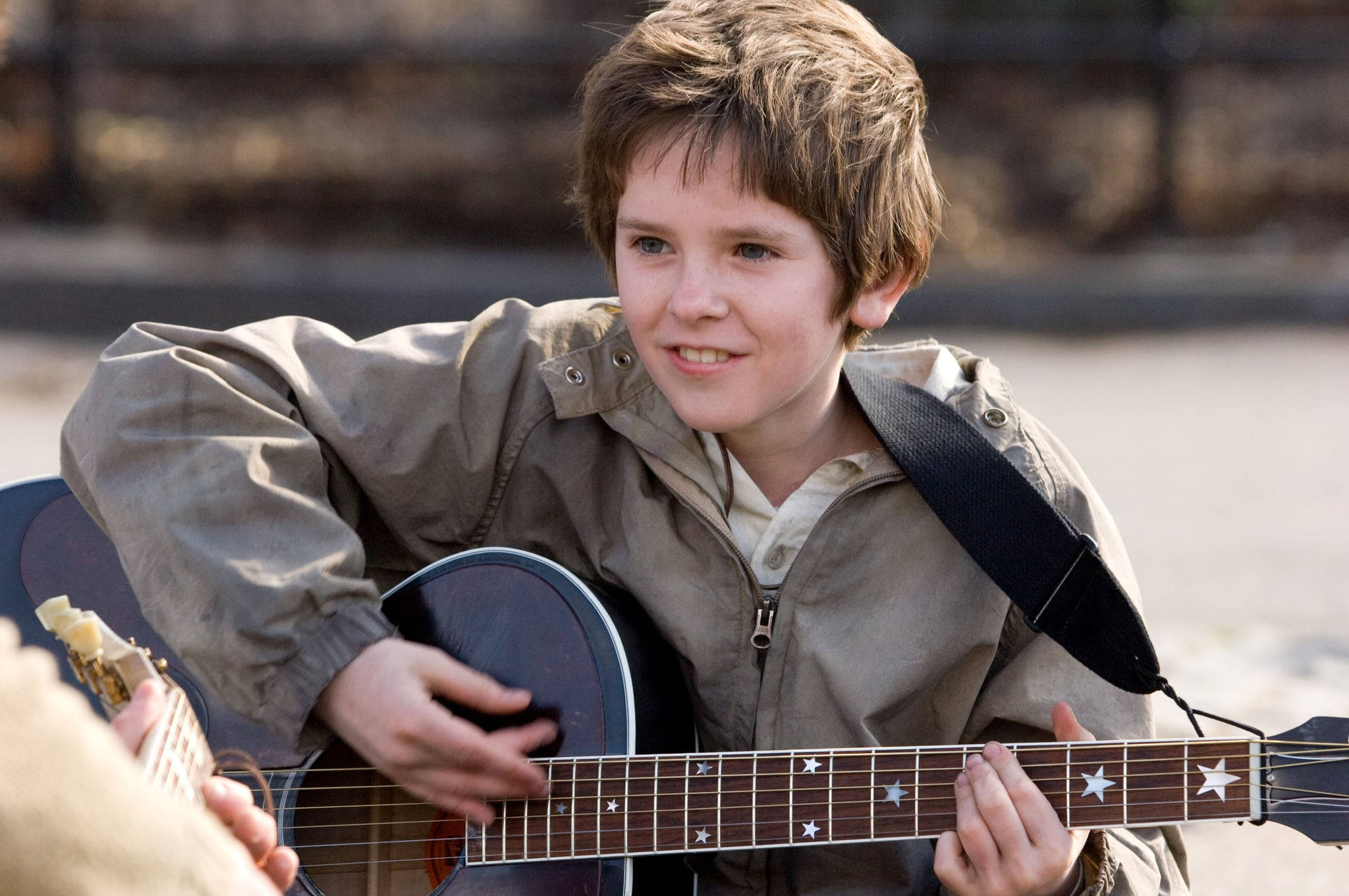 Freddie Highmore 桌布 壁紙 — 一個頭髮蓬鬆的小男孩在戶外彈奏帶有星星鑲嵌的吉他，露出溫暖的笑容。