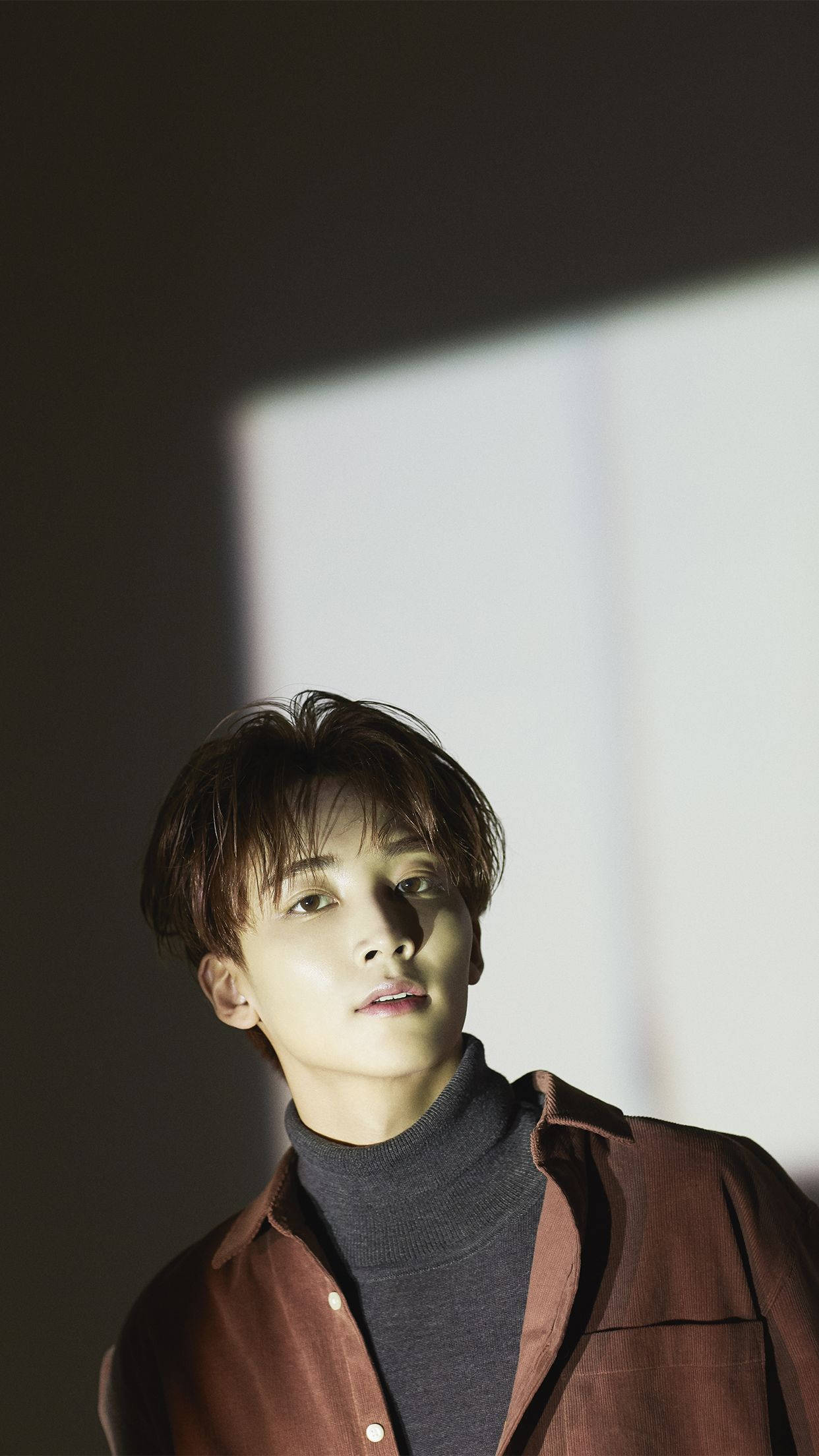 yoon jeonghan 桌布 壁紙 — 一個頭髮蓬鬆的年輕男子在柔和的工作室燈光下向上凝視，一道明亮的矩形光束投射在他的臉和棕色襯衫上。