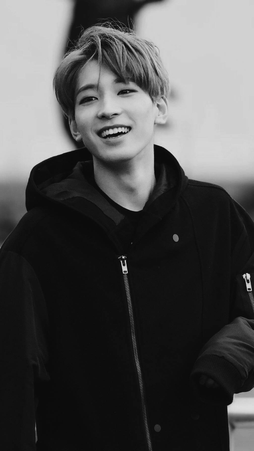Wonwoo 桌布 壁紙 — 一個頭髮蓬鬆的年輕男子穿著黑色連帽衫，在模糊的戶外背景下燦爛地微笑。
