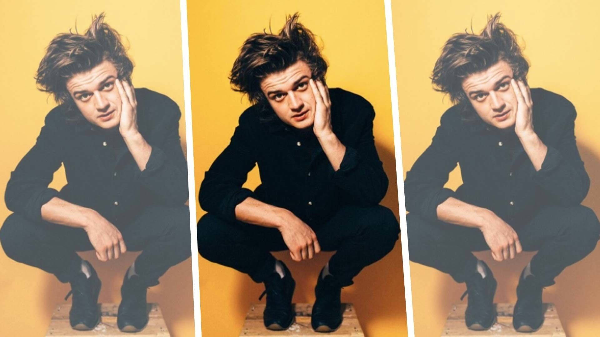 Joe Keery 桌布 壁紙 — 一個頭髮蓬鬆的年輕男子蹲在溫暖的黃色牆邊，手托著臉頰，一副沉思的姿態。
