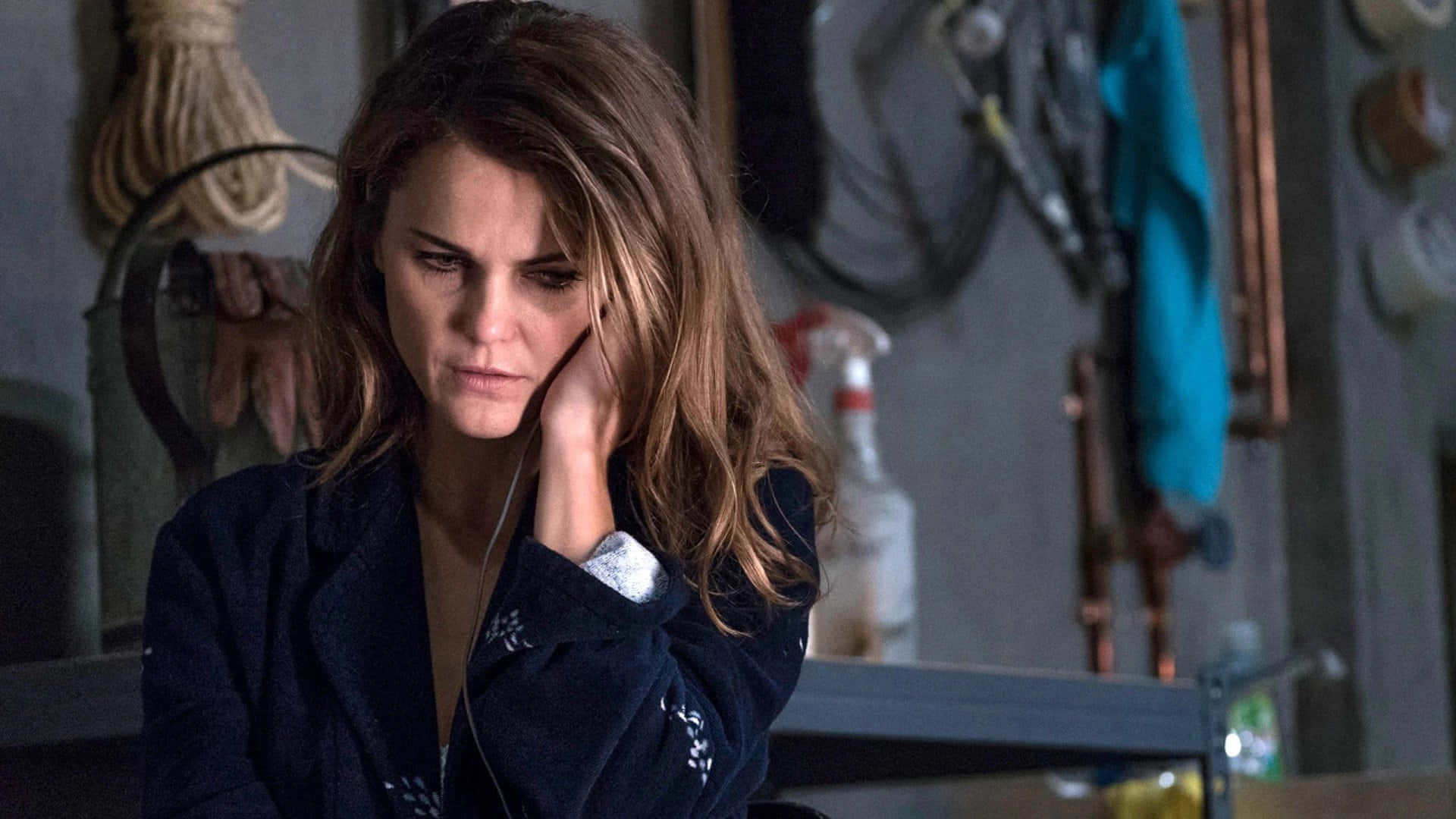 keri russell 桌布 壁紙 — 一個頭髮蓬鬆的棕髮女人在雜亂的車庫裡用手托著頭，顯得疲倦而沉思。