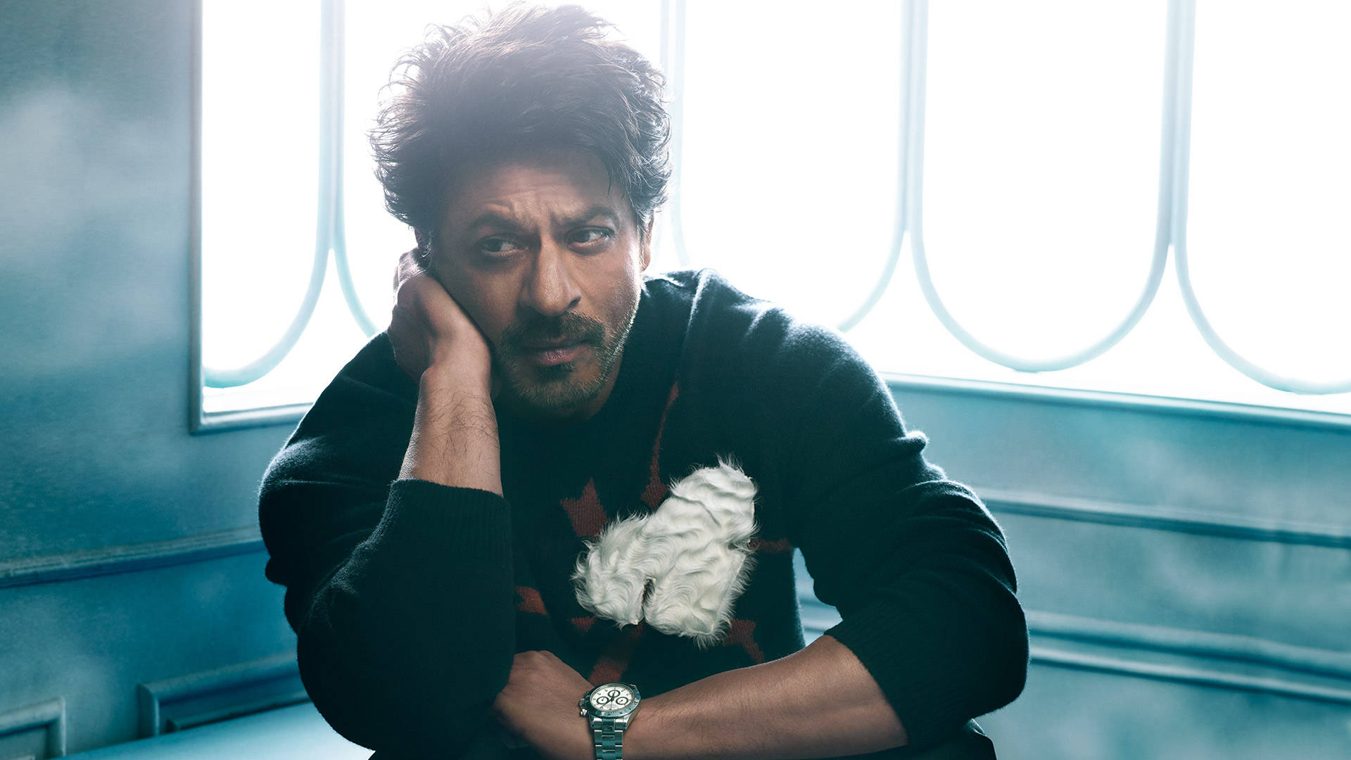 Shahrukh 桌布 壁紙 — 一個頭髮蓬鬆的男人將頭靠在手上，凝視著，穿著黑色毛衣，配白色毛絨飾物，背景是藍色調的窗戶。