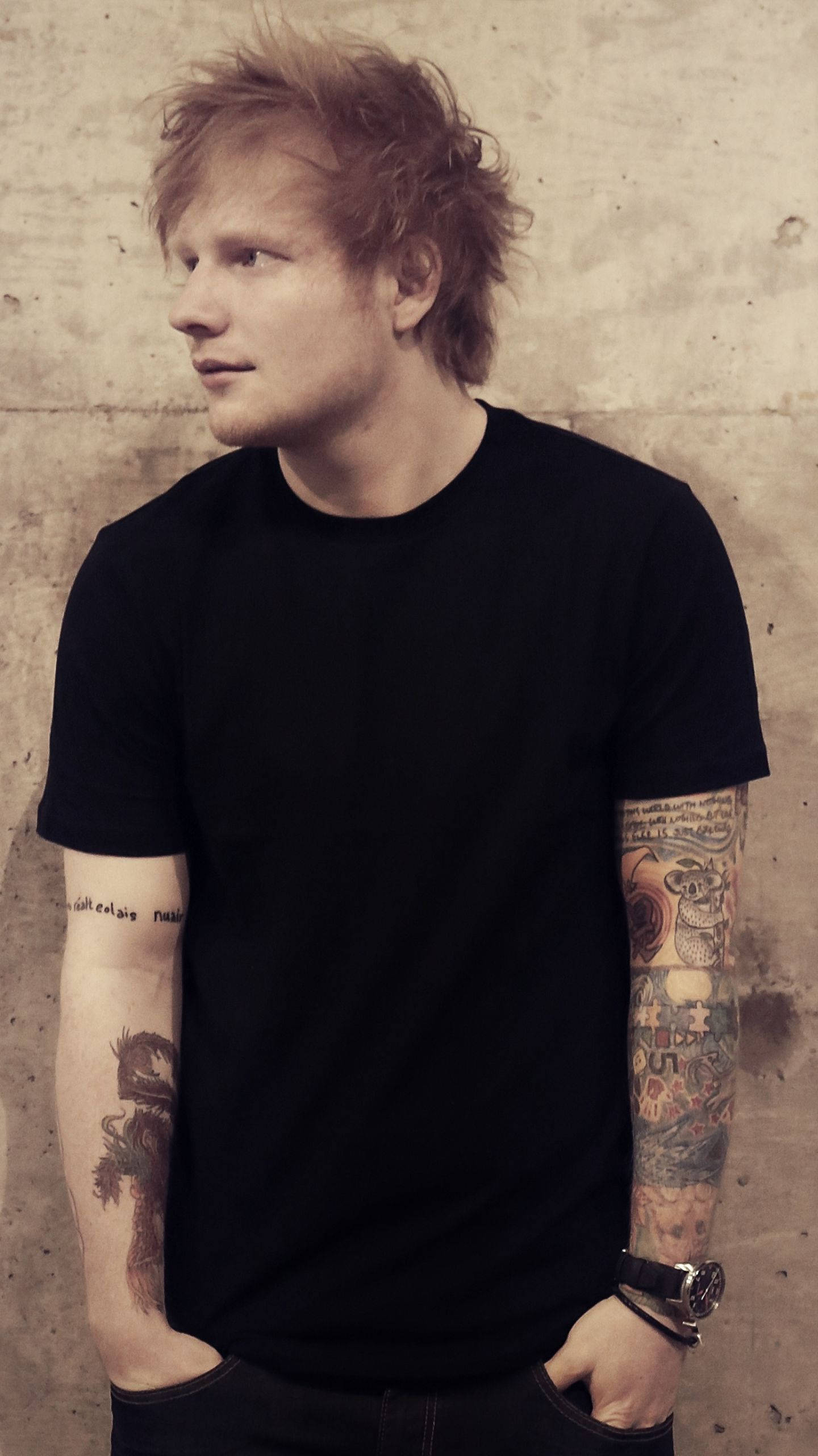 Ed Sheeran 桌布 壁紙 — 一個頭髮蓬鬆的紅髮男子，手臂上有刺青，穿著黑色T恤，靠著紋理混凝土牆站立。