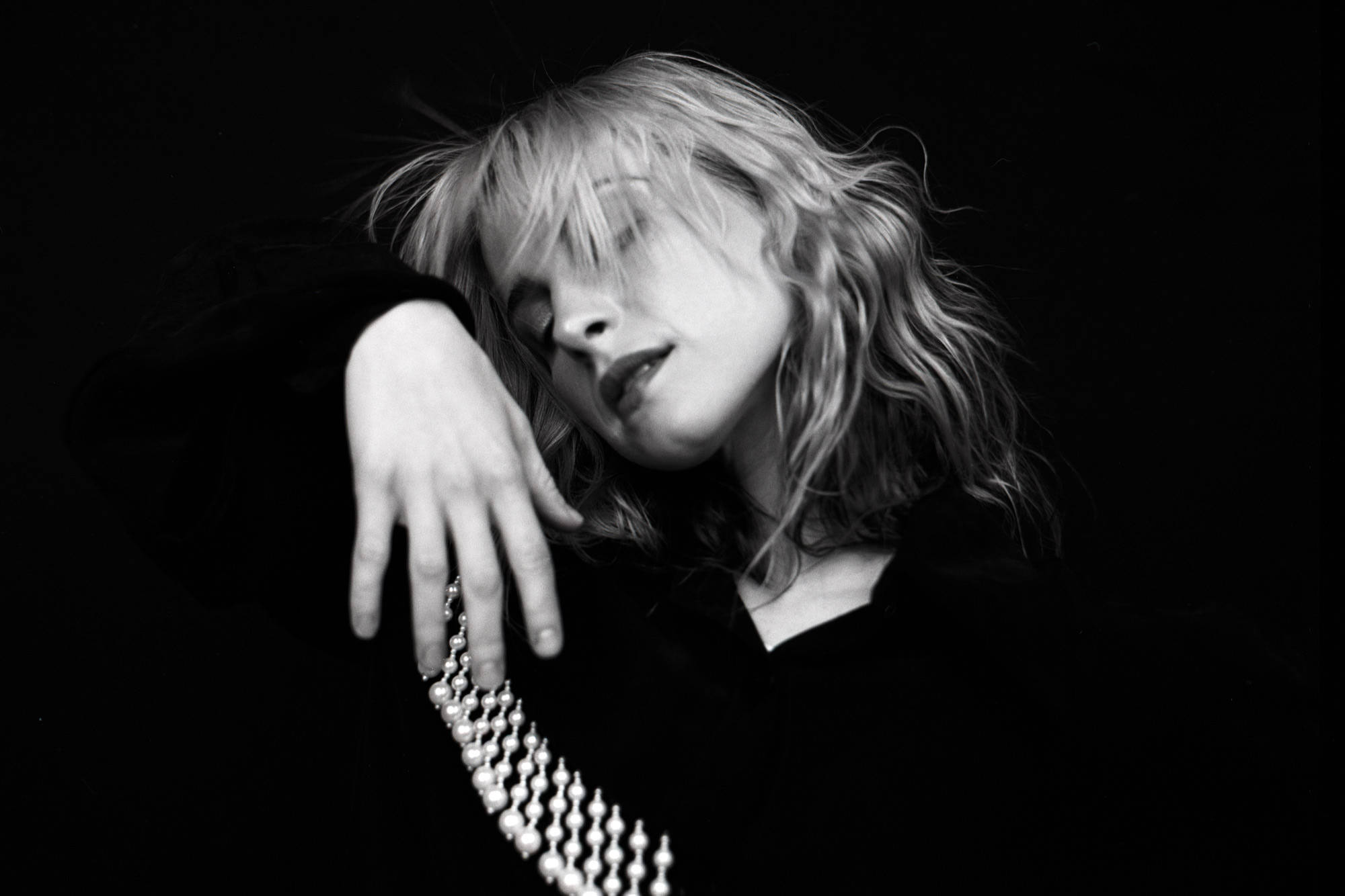 Hayley Williams 桌布 壁紙 — 一個頭髮蓬鬆的金髮女人，戴著珠鏈，頭靠著手，背景是純黑色。