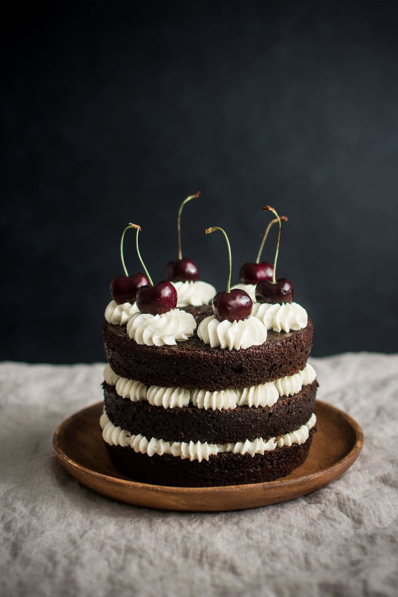 Black Forest Cake 桌布 壁紙 — 一個頹廢的三層巧克力蛋糕，上面有白色奶油糖霜和新鮮櫻桃，優雅地放在木盤上，背景深色。