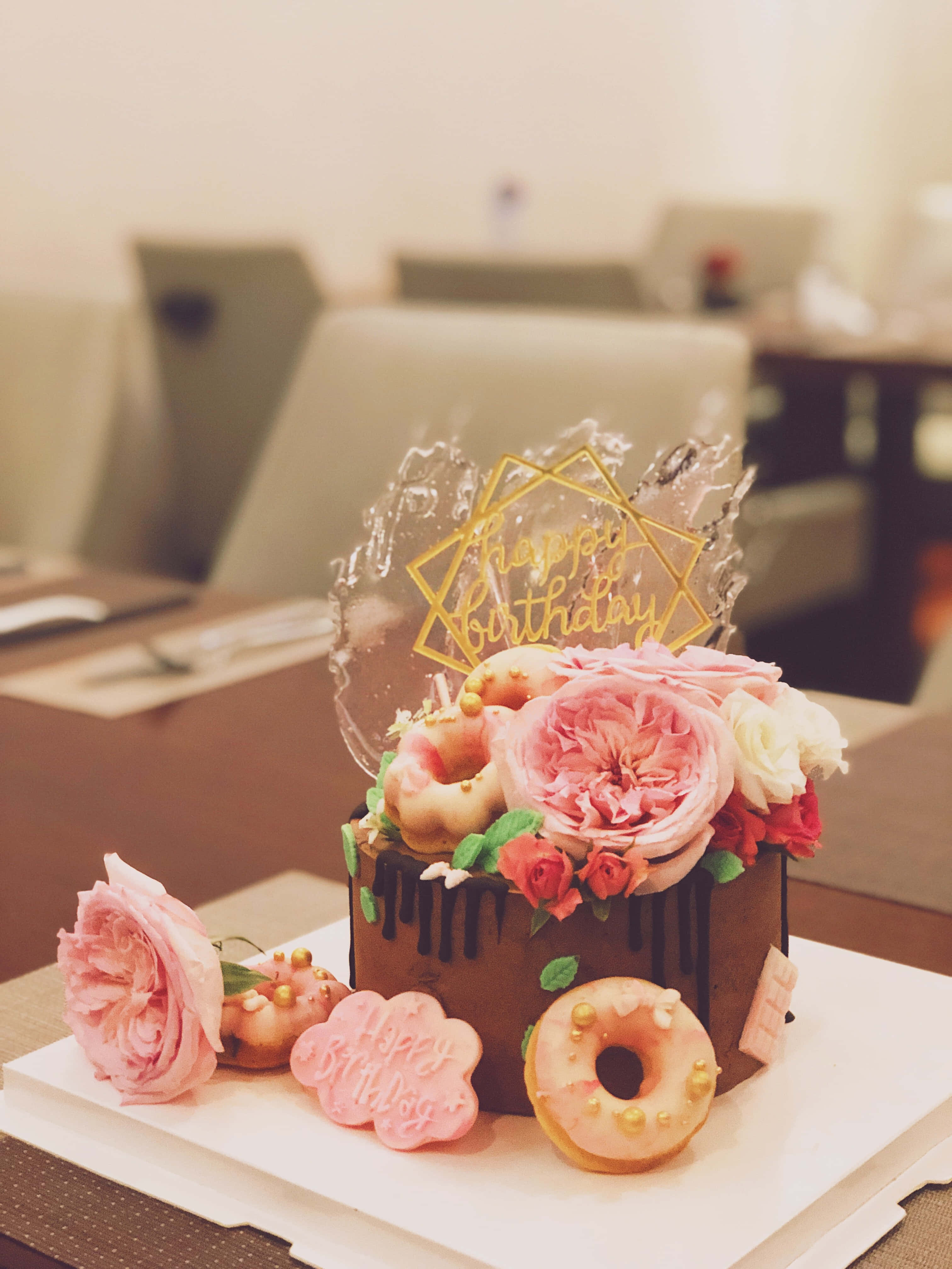 Cute Birthday 桌布 壁紙 — 一個頹廢的巧克力滴落蛋糕，飾有粉紅玫瑰、甜甜圈和金色生日裝飾，優雅地放在白色盤子上。