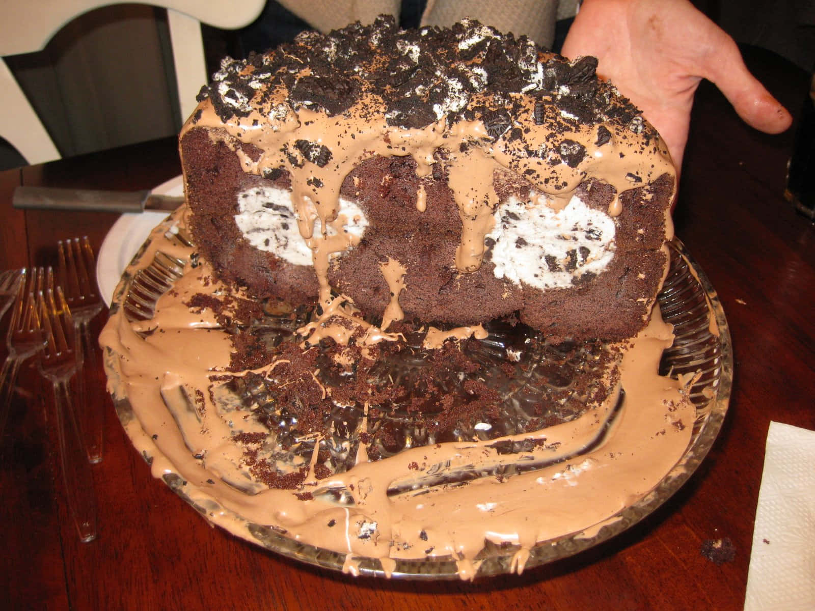 Ugly Cake 桌布 壁紙 — 一個頹廢的巧克力蛋糕，上面有Oreo碎和奶油餡，放在一個凌亂的盤子上，由一隻手在木桌上拿著。