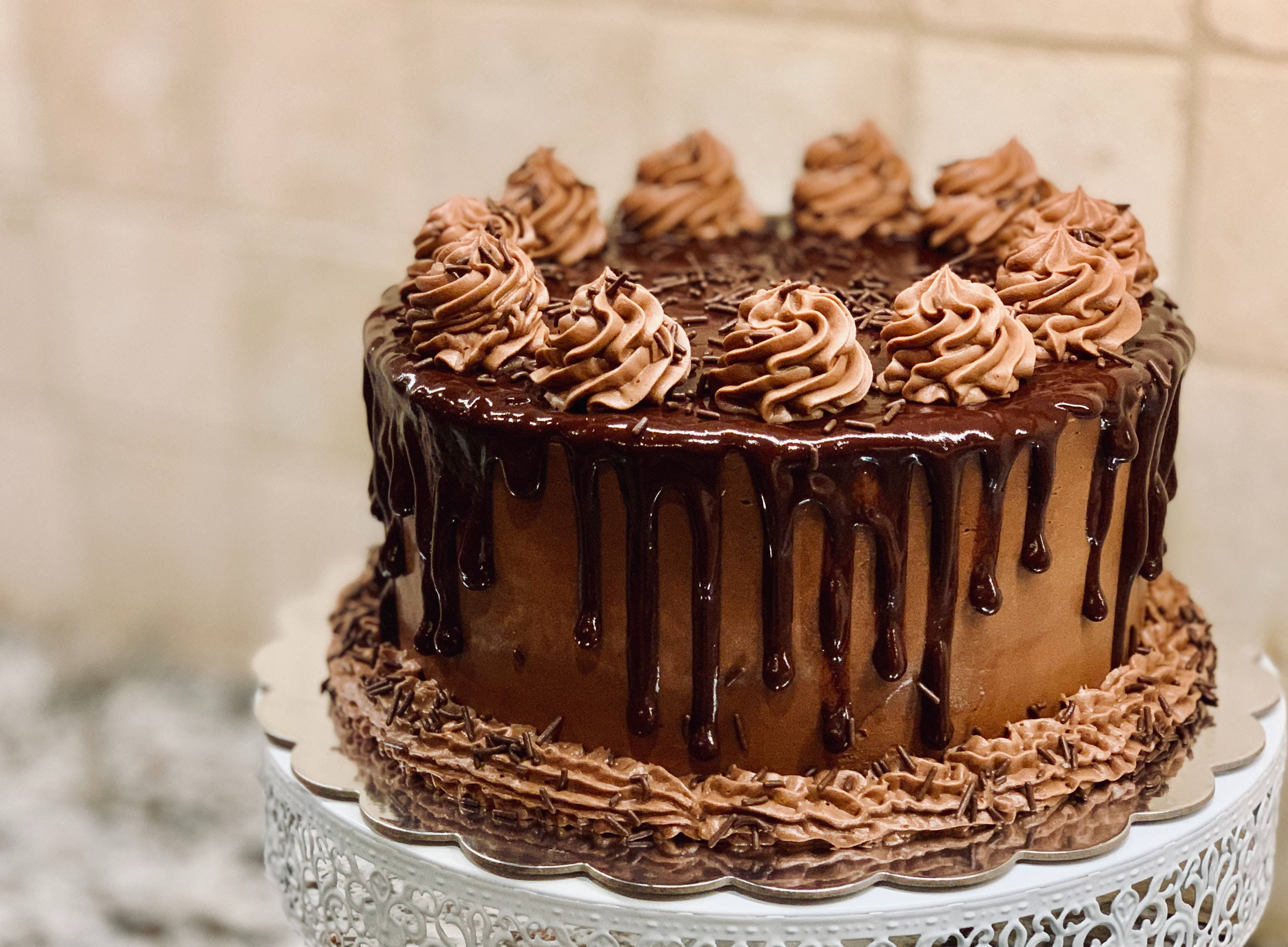 Cake 桌布 壁紙 — 一個頹廢的巧克力蛋糕，帶有光澤的甘納許滴落和擠花漩渦，優雅地放在白色蕾絲蛋糕架上。