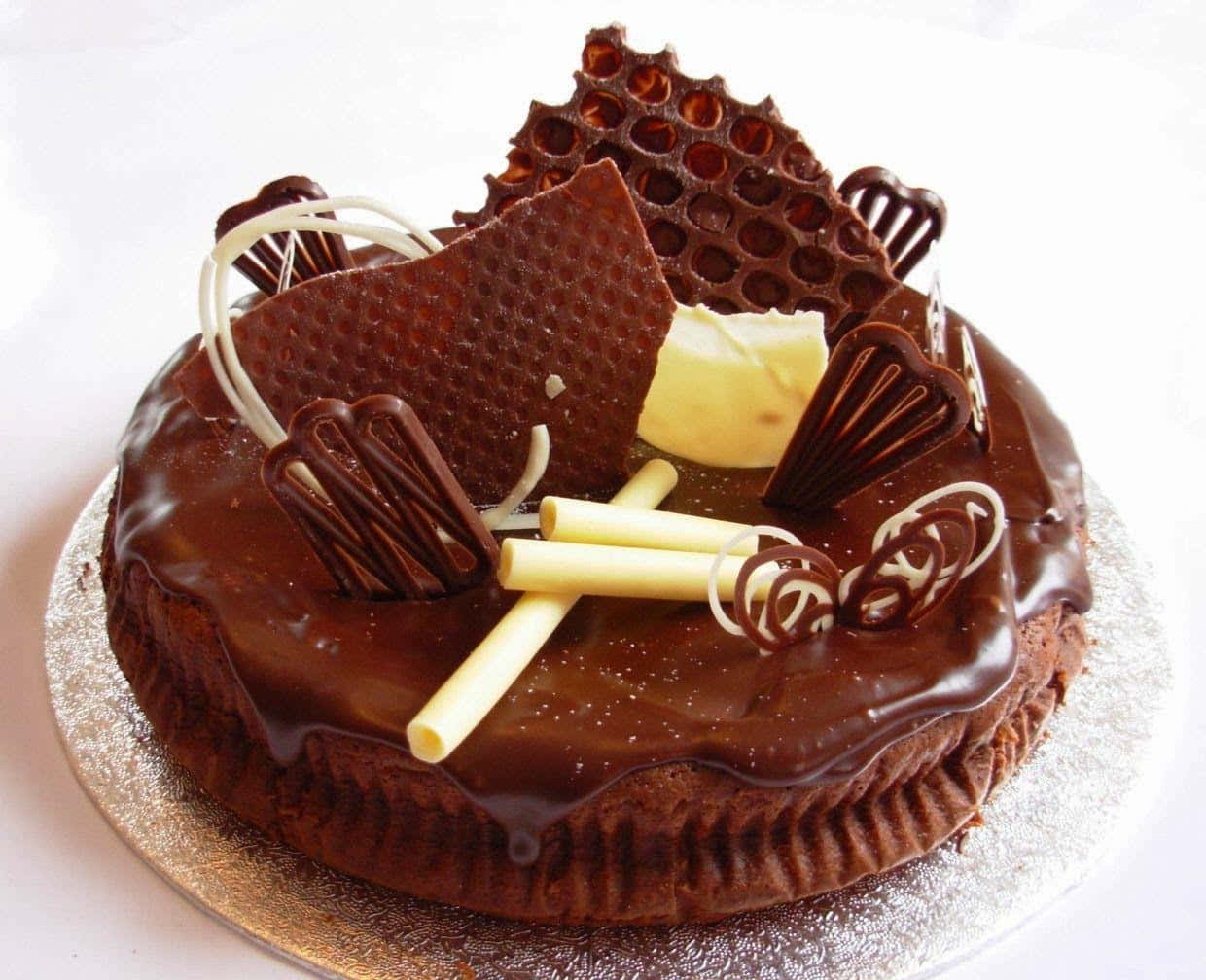 Chocolate Cake 桌布 壁紙 — 一個頹廢的巧克力蛋糕，飾以閃亮的甘納許、白巧克力捲和質感豐富的巧克力碎片，置於銀盤上。