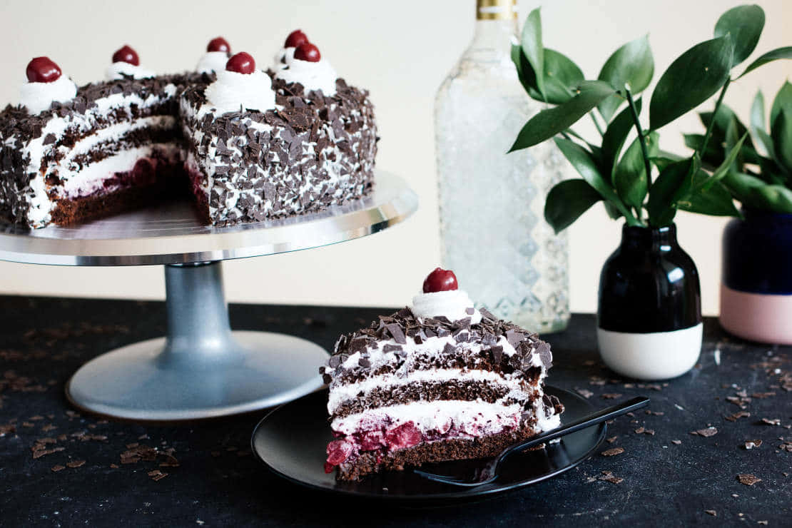 Black Forest Cake 桌布 壁紙 — 一個頹廢的黑森林蛋糕，上面有櫻桃配料，放在銀色架上，旁邊是一片放在黑盤上的蛋糕，撒有巧克力碎屑。