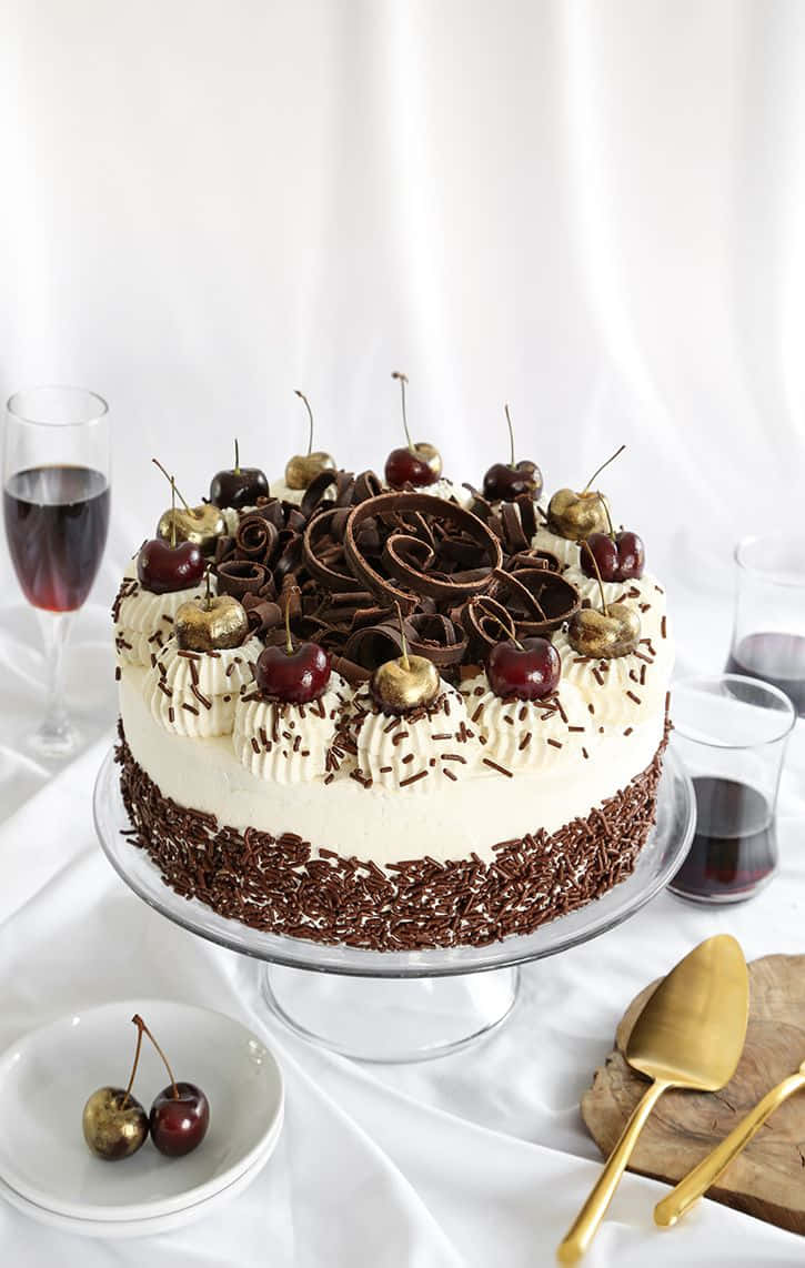 Black Forest Cake 桌布 壁紙 — 一個頹廢的黑森林蛋糕，飾有巧克力捲和光亮的櫻桃，優雅地放在玻璃架上，旁邊是酒杯。