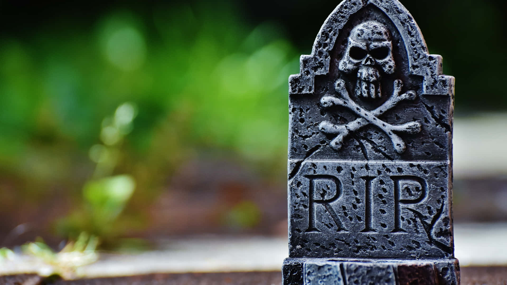 Gravestone 桌布 壁紙 — 一個風化的墓碑，帶有骷髏頭和交叉骨頭標誌及RIP銘文，矗立在模糊的綠色花園中。